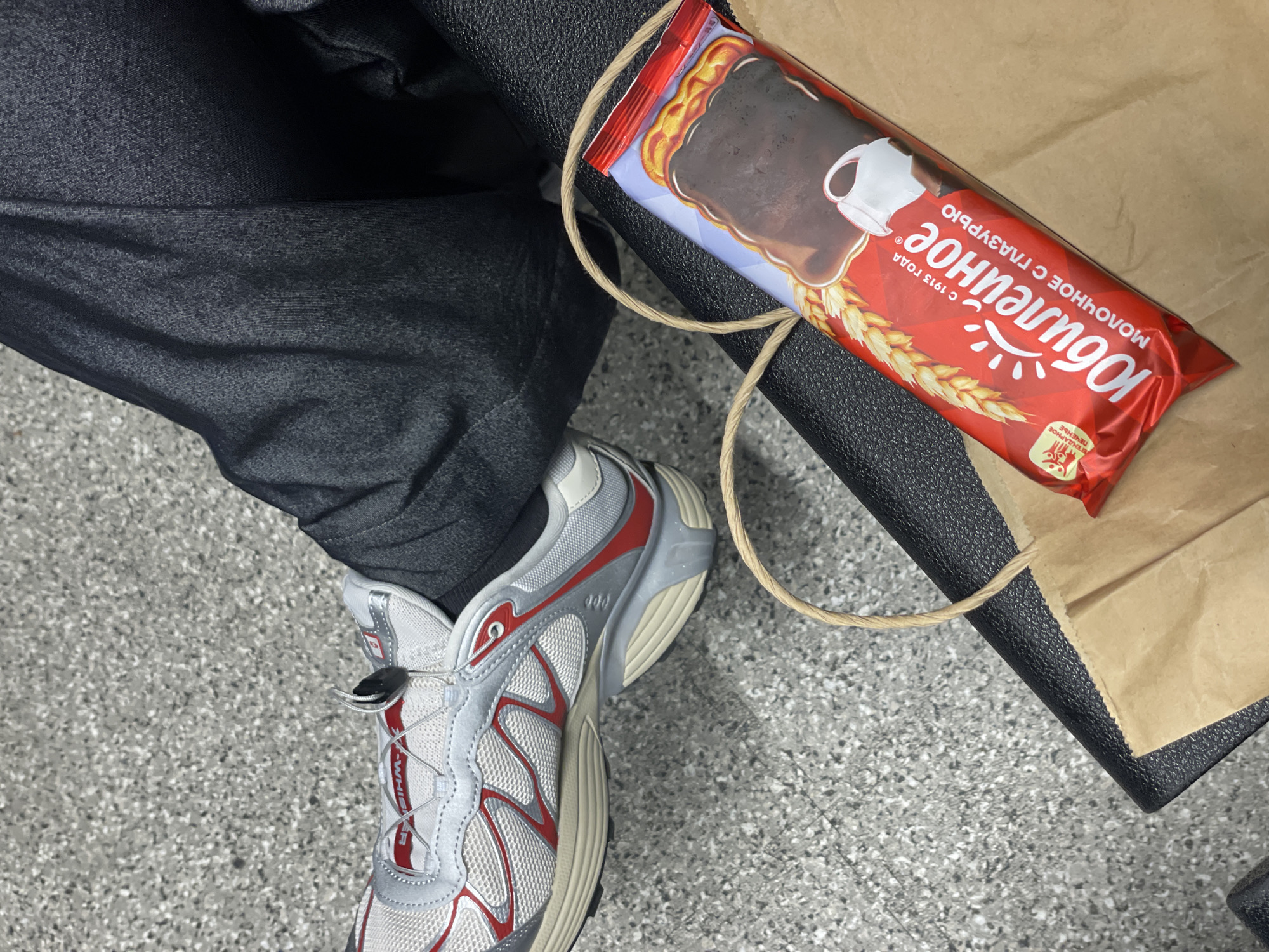 A top-down shot shows a person seated on a black surface. The person wears dark-colored pants and a white, silver, and red sneaker with a toggle lacing system. Beside the person is a brown paper bag containing a red and blue candy bar with wheat graphics, possibly located in Tashkent, Uzbekistan, based on the Cyrillic text on the packaging.