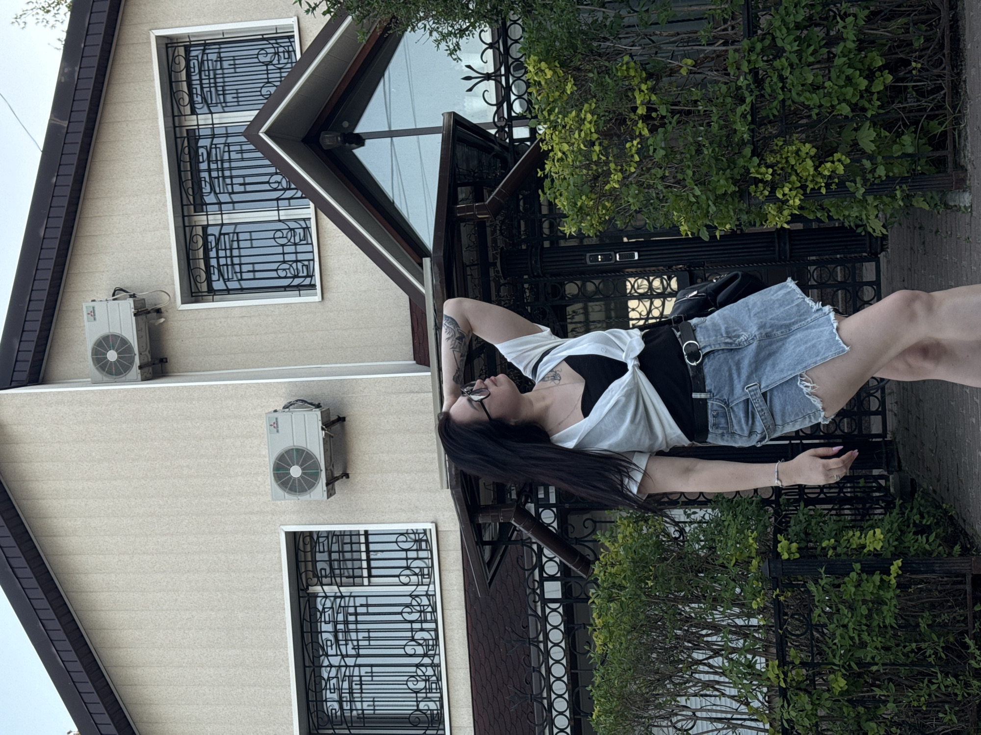A woman with long dark hair, wearing a white shirt over a black top and denim shorts, stands next to a building. The building has two windows with black decorative grilles, and two air conditioning units mounted on the exterior wall. Bushes grow in front of a black metal fence, adding a touch of greenery. The location appears to be an urban environment.