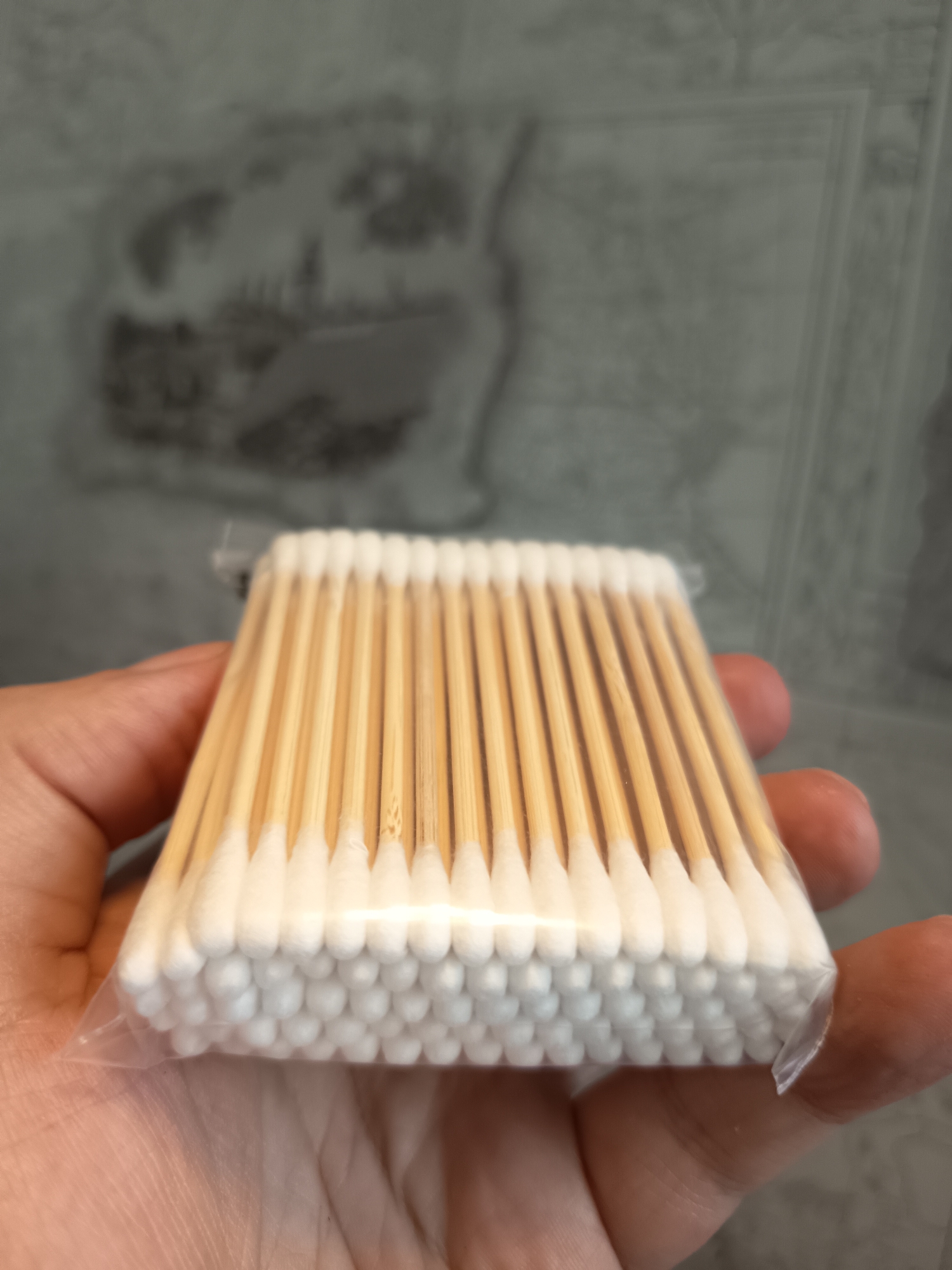 A close-up, eye-level shot shows a person's hand holding a clear plastic package of cotton swabs. The cotton swabs have wooden sticks and white cotton tips. The package is slightly open at the bottom right corner. The background is a blurred, gray, textured surface that appears to be a wall. The image was taken indoors, likely in a bathroom or similar environment. There is no discernible indication of the city of Izmail, Ukraine within the image itself.