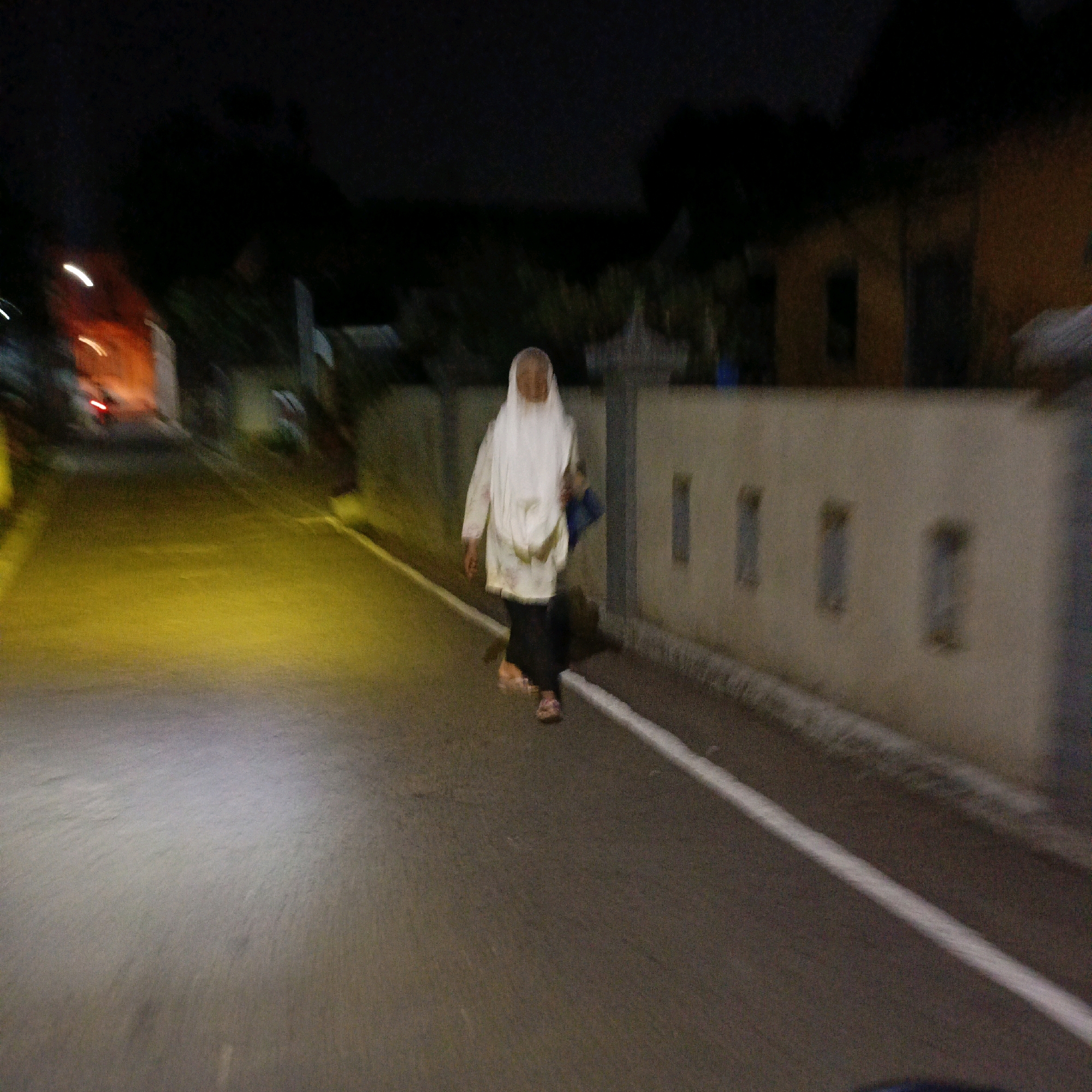 A person wearing a white hijab and a white top over black pants walks on the side of a street at night. The person is carrying a blue bag over their left shoulder. The street has a white line painted on the right side. In the background, a wall with rectangular indentations runs along the street. To the left of the street, there is a bright yellow illuminated area. The scene appears to be in an urban or suburban area, likely in Salatiga, Indonesia, given the context. The lighting suggests it is after dark, and the overall image quality is blurry, indicating movement during capture.