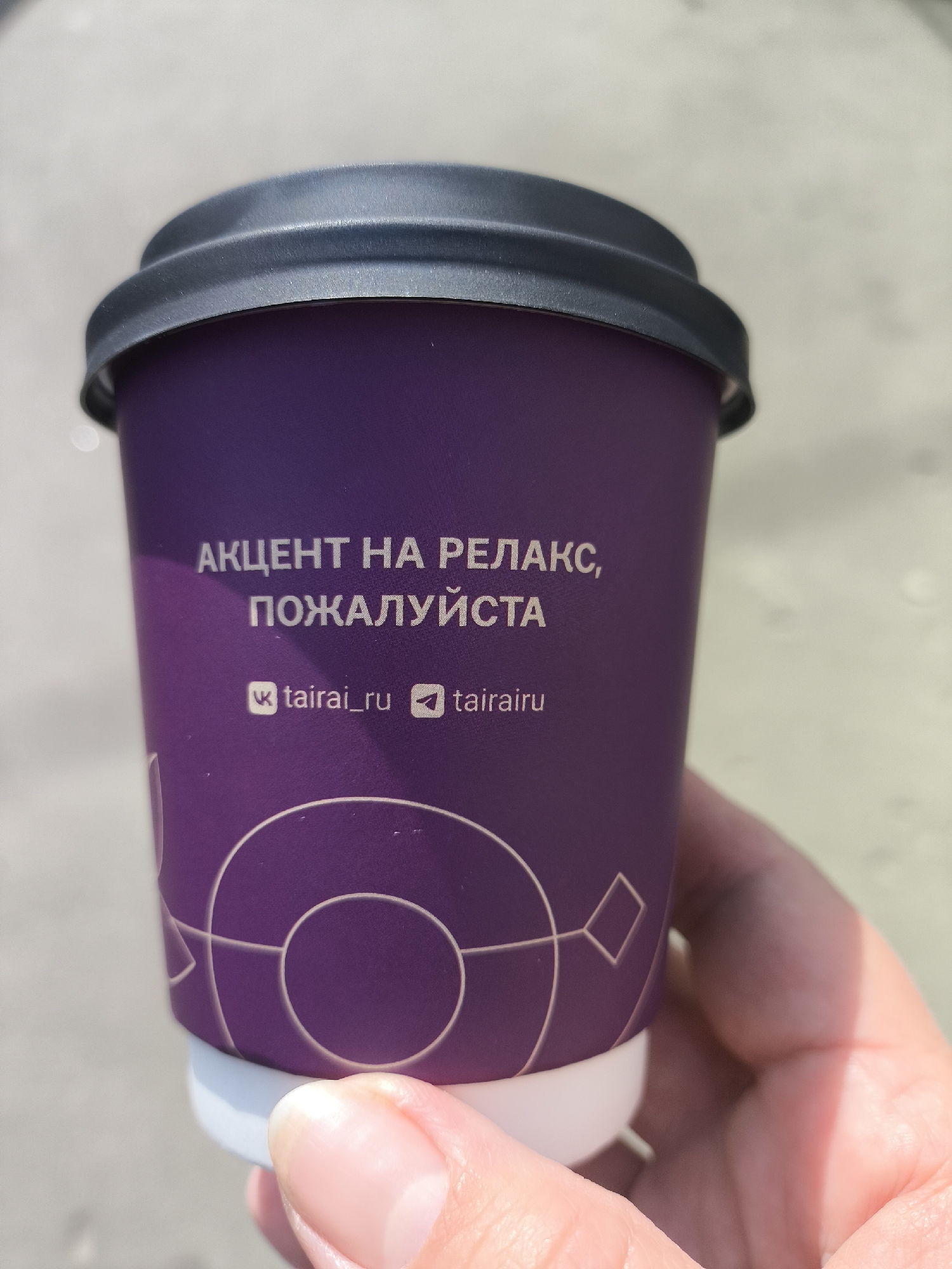 A close-up shot shows a hand holding a purple coffee cup with a black lid. The cup has white text in Russian that reads "АКЦЕНТ НА РЕЛАКС, ПОЖАЛУЙСТА" (Accent on relaxation, please). Below the text are icons and handles: "vk tairai_ru" and "tairairu". A white design circles the base of the cup. The location is likely Moscow, Russia, based on the language of the text.
