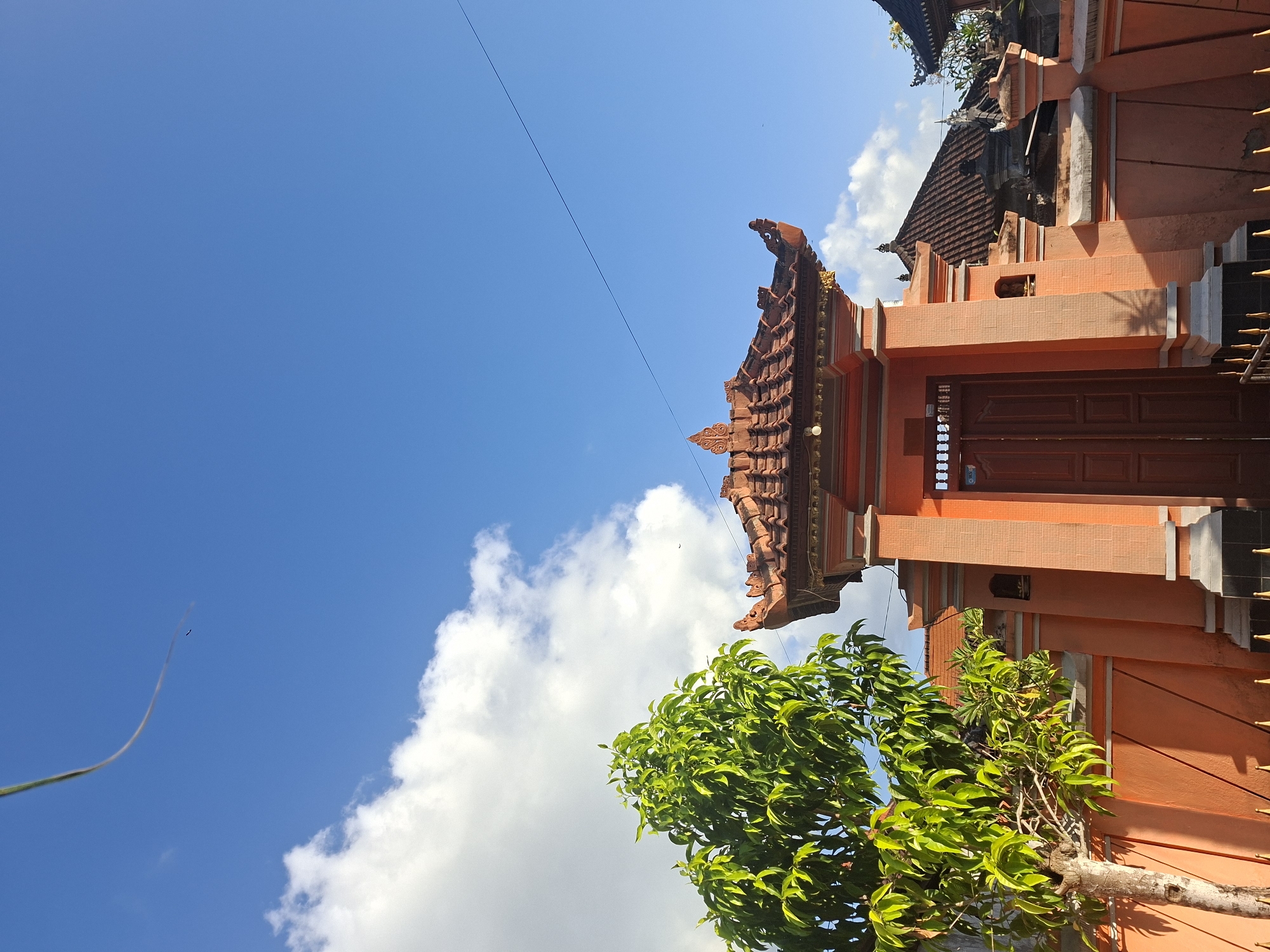 A low-angle, full shot shows an orange building with traditional Balinese architecture, under a bright blue sky with scattered white clouds. The building features ornate, multi-tiered tiled roofs and a central, double-doored entrance. To the left of the entrance, a green tree with lush leaves stands against an orange wall. A thin, dark wire stretches diagonally across the sky from the upper left to the mid-right. The scene appears to be an entrance to a temple or a residential compound in Bali.
