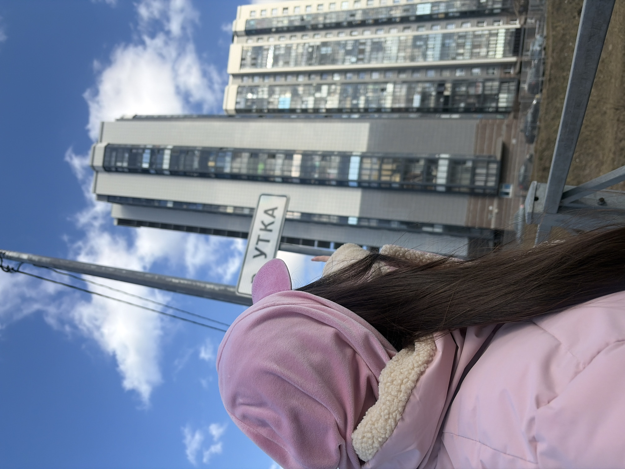 The image depicts an outdoor urban scene in Saint Petersburg, Russia, specifically identifiable by a street sign displaying the partial Cyrillic inscription "УТКА," likely referring to Utkina Ulitsa. In the foreground, a person is observed from a rear-left perspective, facing away from the camera and towards the background. This individual has long, straight dark brown hair and is dressed in a light pink hooded jacket featuring a visible cream-colored sherpa lining along the collar. A matching light pink, possibly velvet, hooded top with a small visor is pulled over their head. Their right hand is partially visible against the jacket, appearing slightly raised.
The background is dominated by two modern high-rise residential buildings under a clear blue sky with scattered white cumulus clouds. The building on the left is a tall structure with a grey facade and numerous dark-tinted glass windows and balconies. The building on the right is also a multi-story residential complex, slightly lighter in tone, with a facade featuring numerous windows and balconies. A tall, dark grey utility pole with wires extends vertically through the image, partially obscuring the left building, and supports the white rectangular "УТКА" street sign. Below the buildings, a dry, brown ground embankment is visible, with an out-of-focus road and several dark-colored vehicles in the distance. A grey metal railing runs horizontally across the lower foreground, suggesting an elevated vantage point.