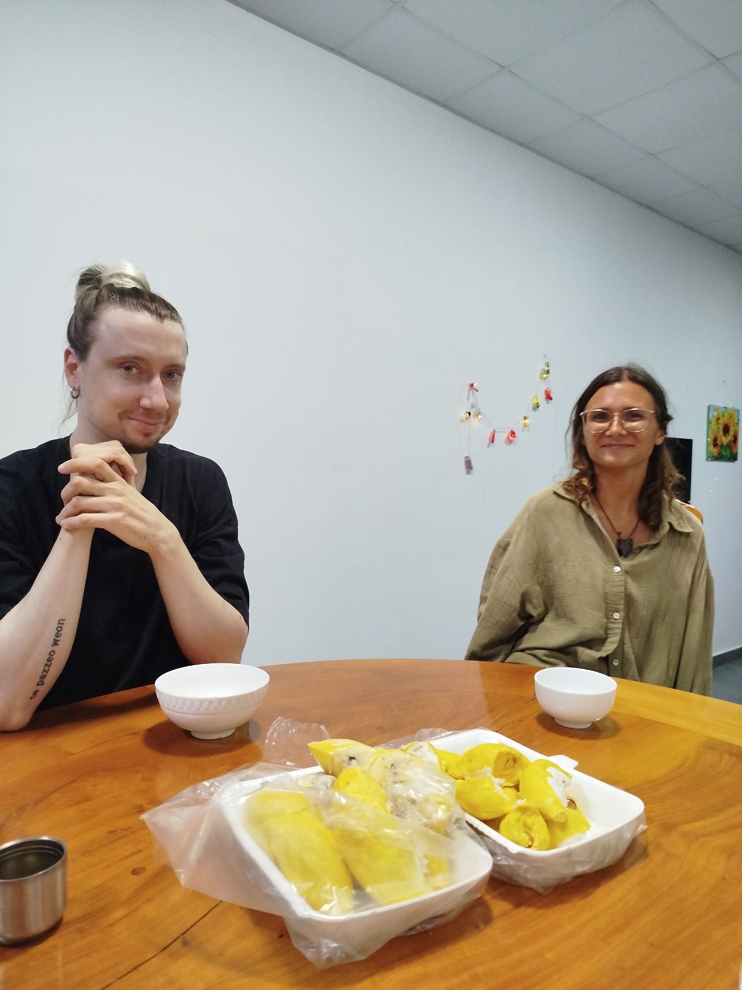 Two people, a man and a woman, are seated at a round wooden table indoors. On the table are two white bowls, a metal cup, and two white containers filled with yellow fruit, possibly durian, partially covered with plastic wrap. The man has long blond hair in a bun and a tattoo on his forearm. He is wearing a black shirt and has his hands clasped. The woman wears glasses and an olive-green button-down shirt. Behind the woman, the wall is decorated with string lights and a framed picture of sunflowers. This scene is located in the city of Da Lat, Vietnam.