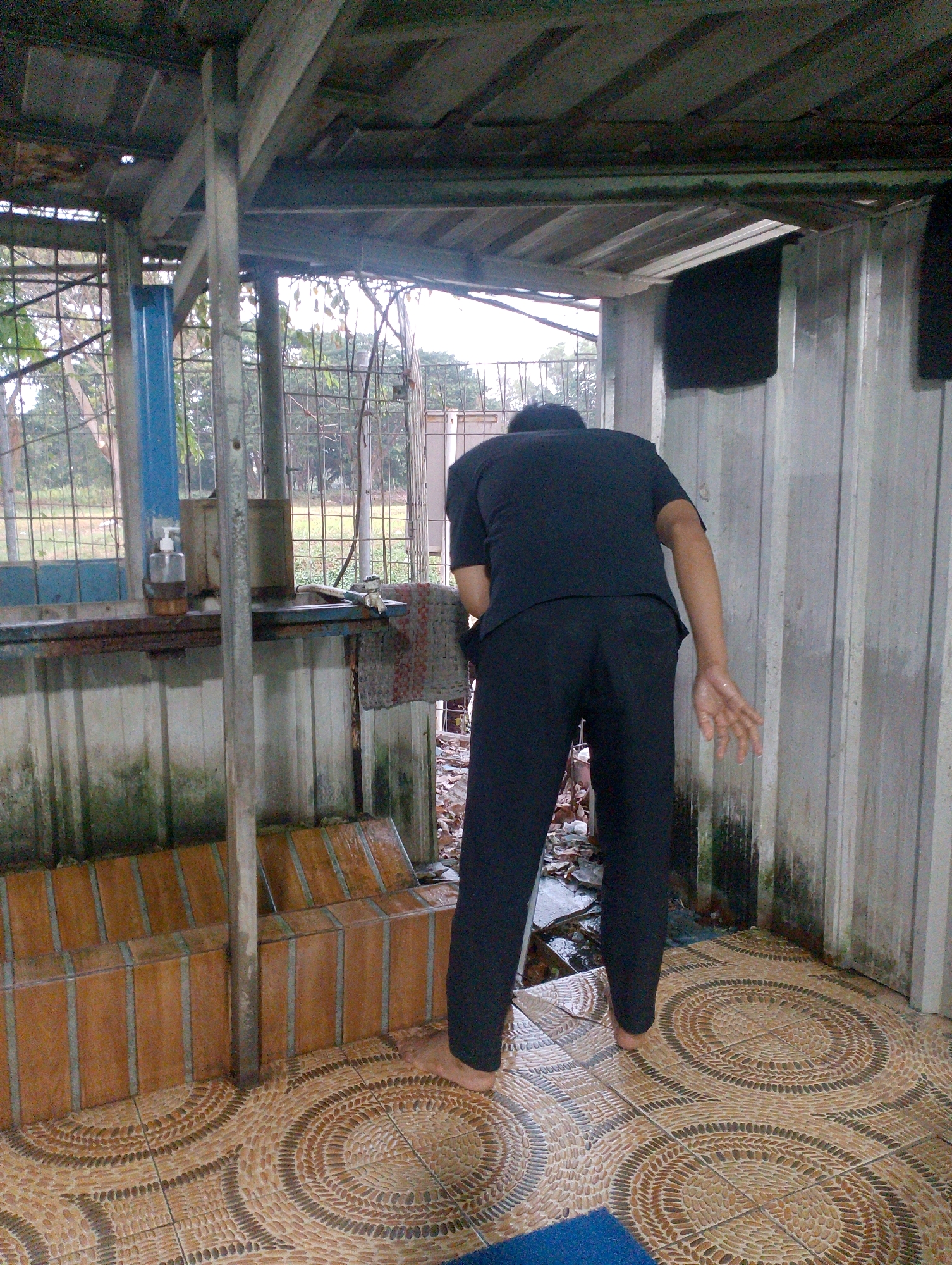 The image shows a person from behind, wearing a black t-shirt and black pants, bending over. The person is standing on a tiled floor with a circular pattern. To the left, there is a raised tiled structure, possibly a bench, and a blue vertical pole. Behind the bench, there is a metal fence with vegetation visible beyond it. The structure appears to be outdoors and is partially covered by a corrugated metal roof supported by metal beams. The walls are also made of corrugated metal. A hand soap dispenser is visible on a shelf. The scene is located in Tangerang, Indonesia, likely a public restroom or ablution area.