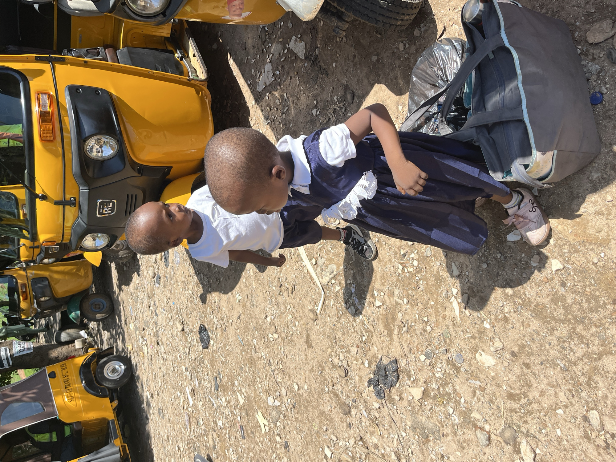 The scene depicts an outdoor environment in Galadimawa Village, Nigeria, characterized by an unpaved, dusty ground with scattered debris. Two young children are visible in the foreground. A boy, approximately 3-5 years old, stands center-left, wearing a white short-sleeved t-shirt, dark shorts, and black and white sneakers. He has short dark hair and is looking towards the right. A girl, of similar age, stands center-right, wearing a dark blue dress with a white collar and white lace detailing, light pink shoes, and white socks. She also has short dark hair and is looking towards the left. Neither child appears to be directly interacting with the other or any specific object. In the background, several yellow auto-rickshaws (tuk-tuks) are parked. One on the far left has "SAJDAN TRANS SERVICES" written on its side. A prominent yellow auto-rickshaw in the mid-ground displays the "BAJAJ RE" logo on its front. Part of another yellow auto-rickshaw is visible on the far right. The ground is littered with small stones, pieces of plastic, and other refuse. Near the girl's feet, a large dark gray tote bag with light blue trim sits on the ground, and a black plastic bag is positioned behind it. Shadows cast on the ground indicate bright sunlight.