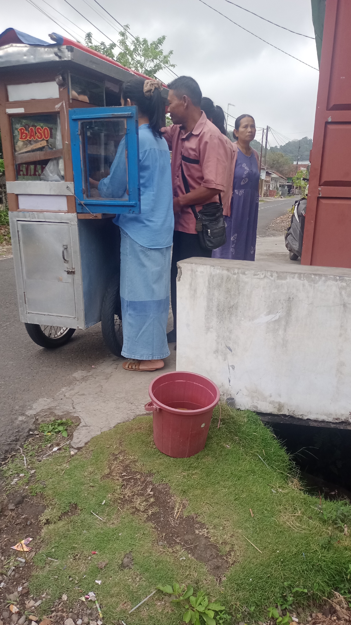 A man in a pink collared shirt and a woman in a blue long-sleeved shirt are standing by a food cart labeled "BASO." The man is looking at the woman and appears to be speaking to her. The woman is wearing a long blue skirt and appears to be working at the food cart. A second woman in a purple dress is standing behind the man, looking towards the right. A red bucket filled with liquid sits on the grass next to a concrete barrier. The food cart is on a paved road. The scene is likely in Pakemitan, Indonesia, given the context of the food cart and the appearance of the people. The sky is overcast.