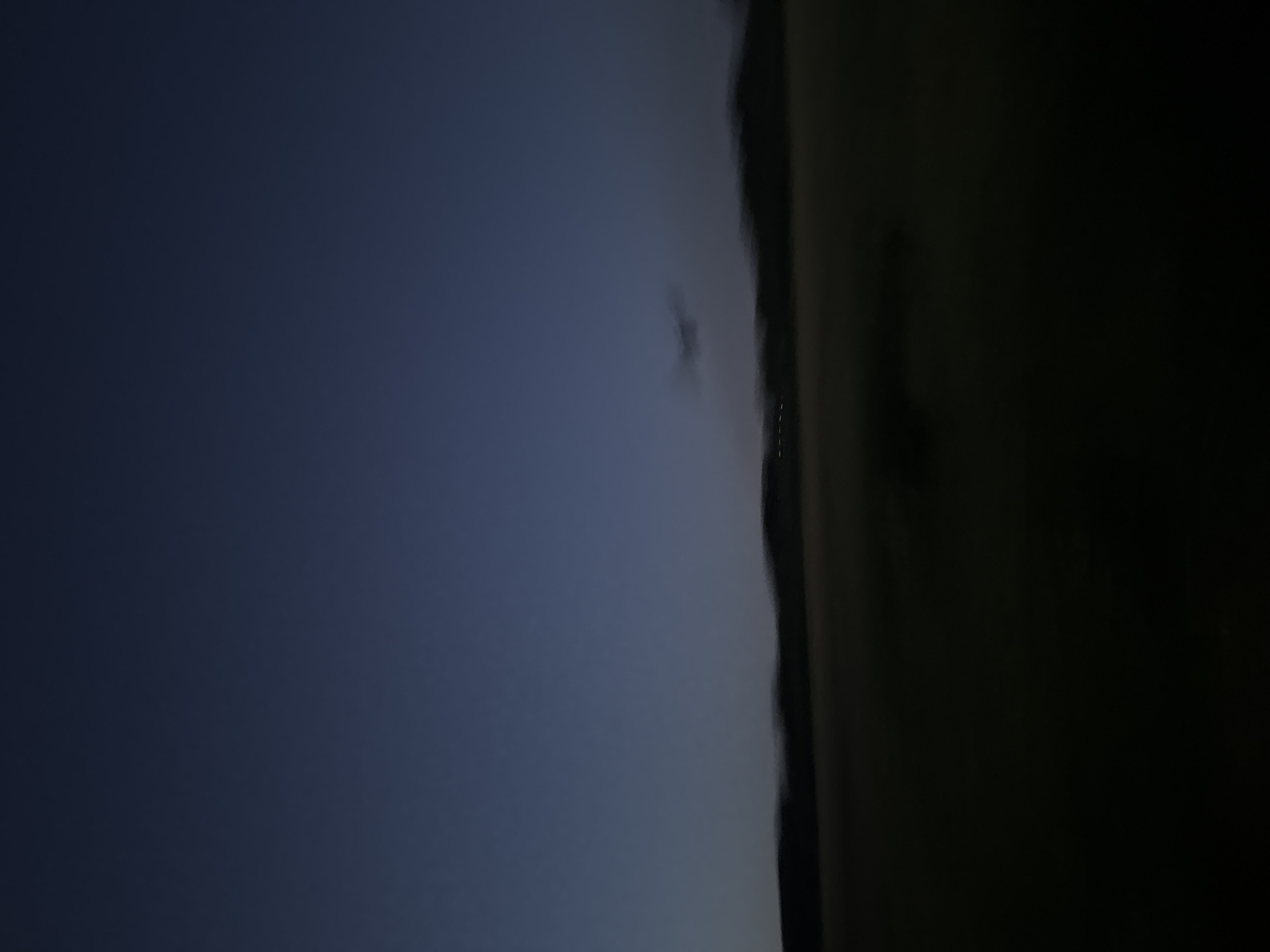 The image captures a blurry nighttime scene with a dark blue sky above a dark, undulating landscape. In the distance, a faint line of lights suggests a settlement. A faint, indistinct object, possibly a cloud formation or a distant aircraft, is visible against the sky. The overall impression is one of a rural or semi-rural area under a twilight or early night sky, characteristic of the outskirts of Pavlodar, Kazakhstan.