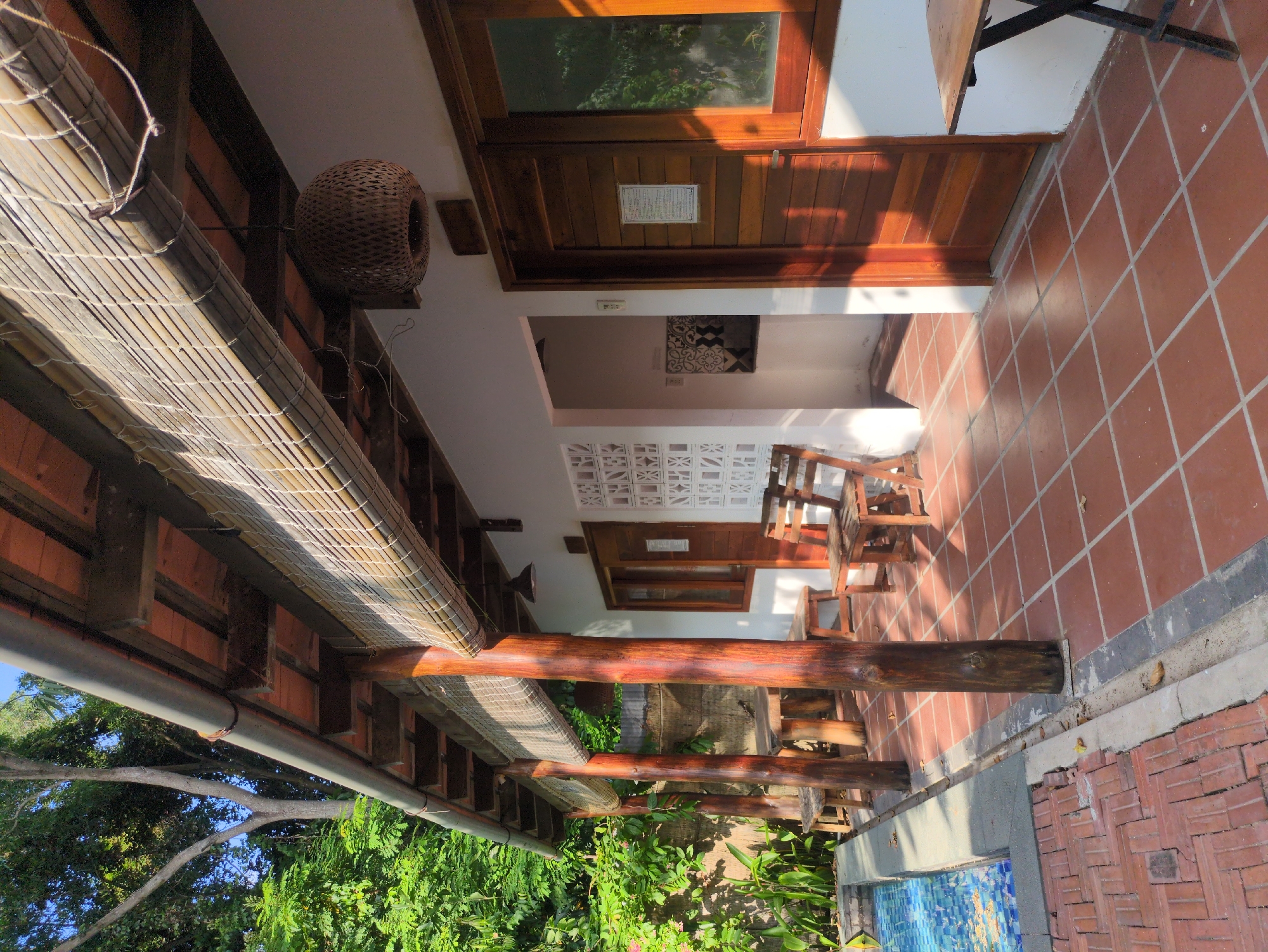 The image captures an outdoor space in Phú Quốc, Vietnam. The foreground consists of a red brick-tiled terrace and seating area. Wooden chairs and tables are arranged in a line, suggesting a restaurant or cafe. Overhanging is a traditional Vietnamese roof structure with a bamboo shade, providing protection from the sun. Lush greenery, including trees and shrubs, is visible below the roof. To the left, the scene extends to a view of the sky. The overall setting has an authentic, local Vietnamese aesthetic.