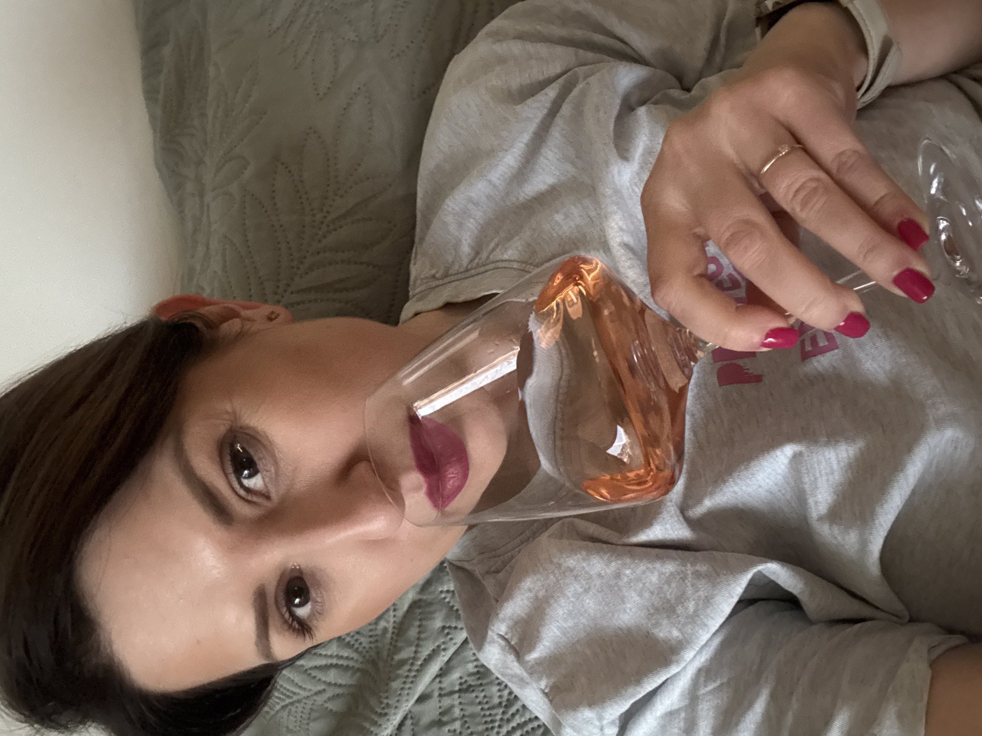 A person is reclining on a bed with a quilted gray cover. The person is wearing a gray t-shirt and holding a wine glass filled with a peach-colored liquid. The person has brown hair, brown eyes, and is wearing dark lipstick. The person is also wearing a ring on their right hand, and their nails are painted a dark pink. The location cannot be determined from the image, and there is no indication the image was taken in Sofia, Bulgaria.