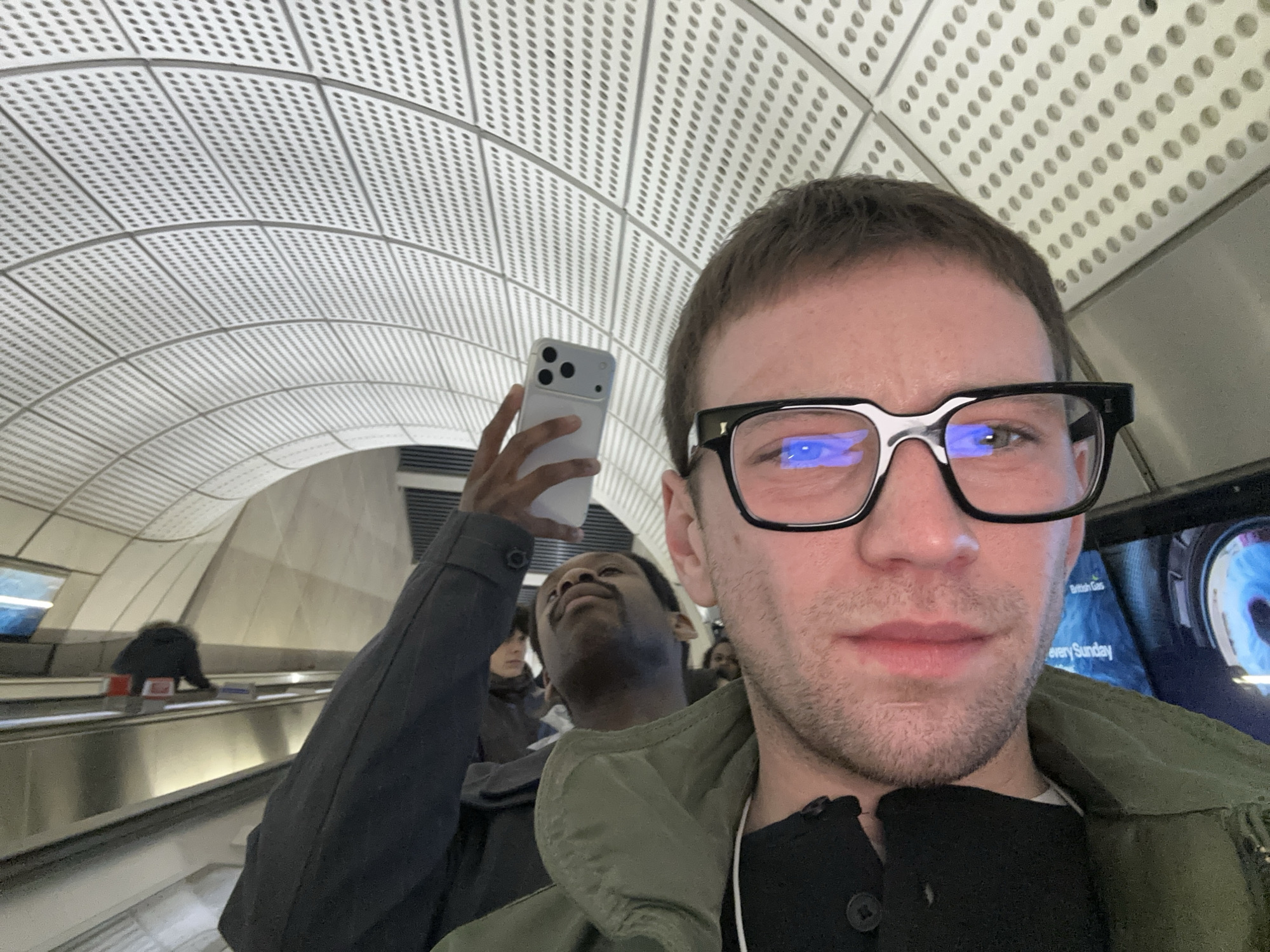 A first-person, close-up view shows a man wearing eyeglasses in the foreground, with another man holding a smartphone behind him. Both are on an escalator. The background shows the curving, perforated ceiling characteristic of a London Underground station. Other individuals can be seen on the adjacent escalator. The advertisement on the display indicates the British Gas company. This photograph was taken in the London Underground.