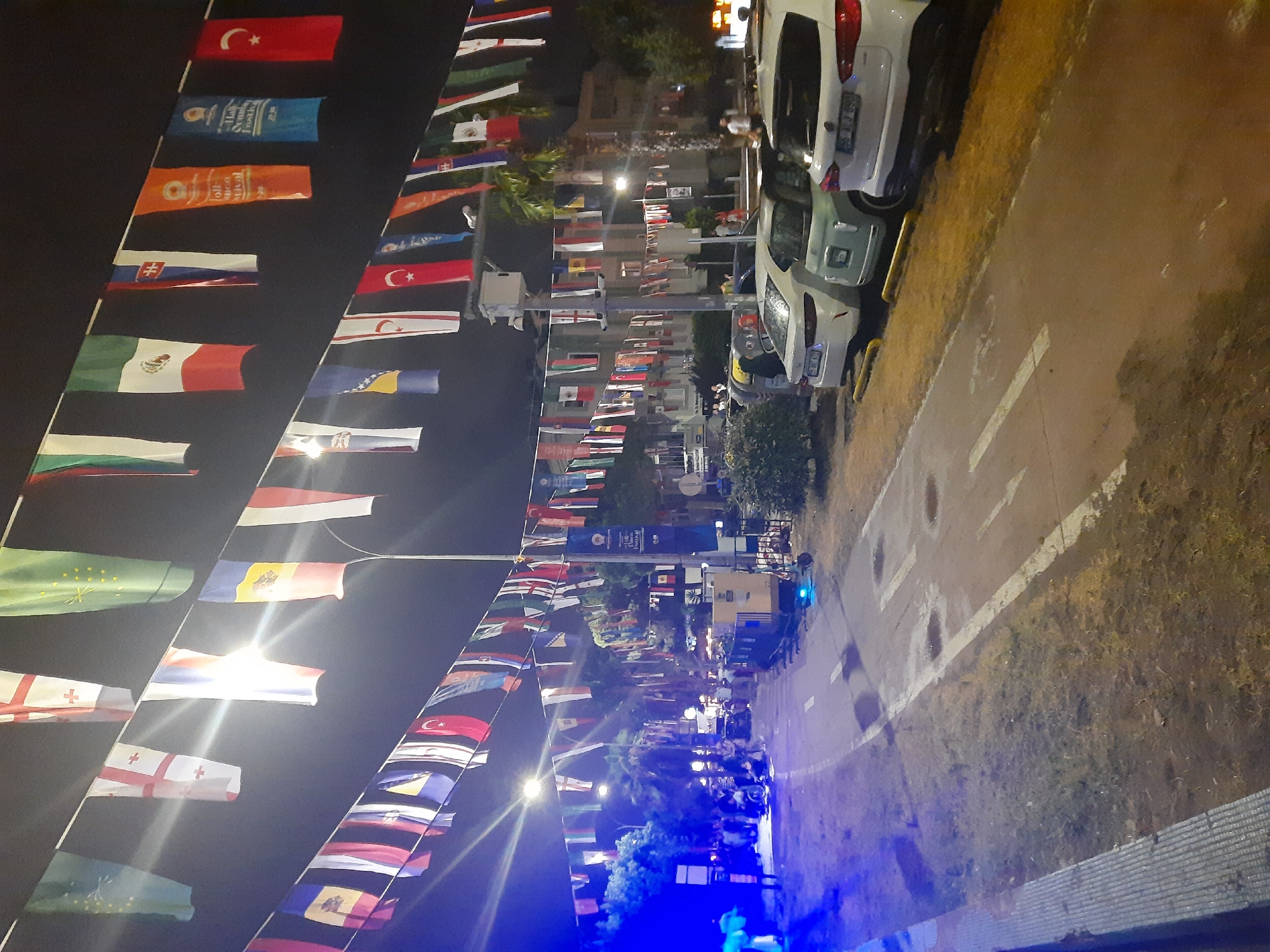 The image depicts a street scene at night in Atakum, Turkey, adorned with numerous international flags suspended across the street on wires. A pathway, possibly a pedestrian or bicycle lane, curves through the foreground, with white lines marking its edges and direction. Several cars are parked along the right side of the pathway, including white SUVs and a light blue compact car. In the background, a crowd of people is visible on the left side of the street, some illuminated by blue lights, suggesting an event or gathering. Buildings and trees line the street, with streetlights providing illumination. The overall atmosphere is festive, likely due to the display of flags, hinting at a cultural festival or international event.