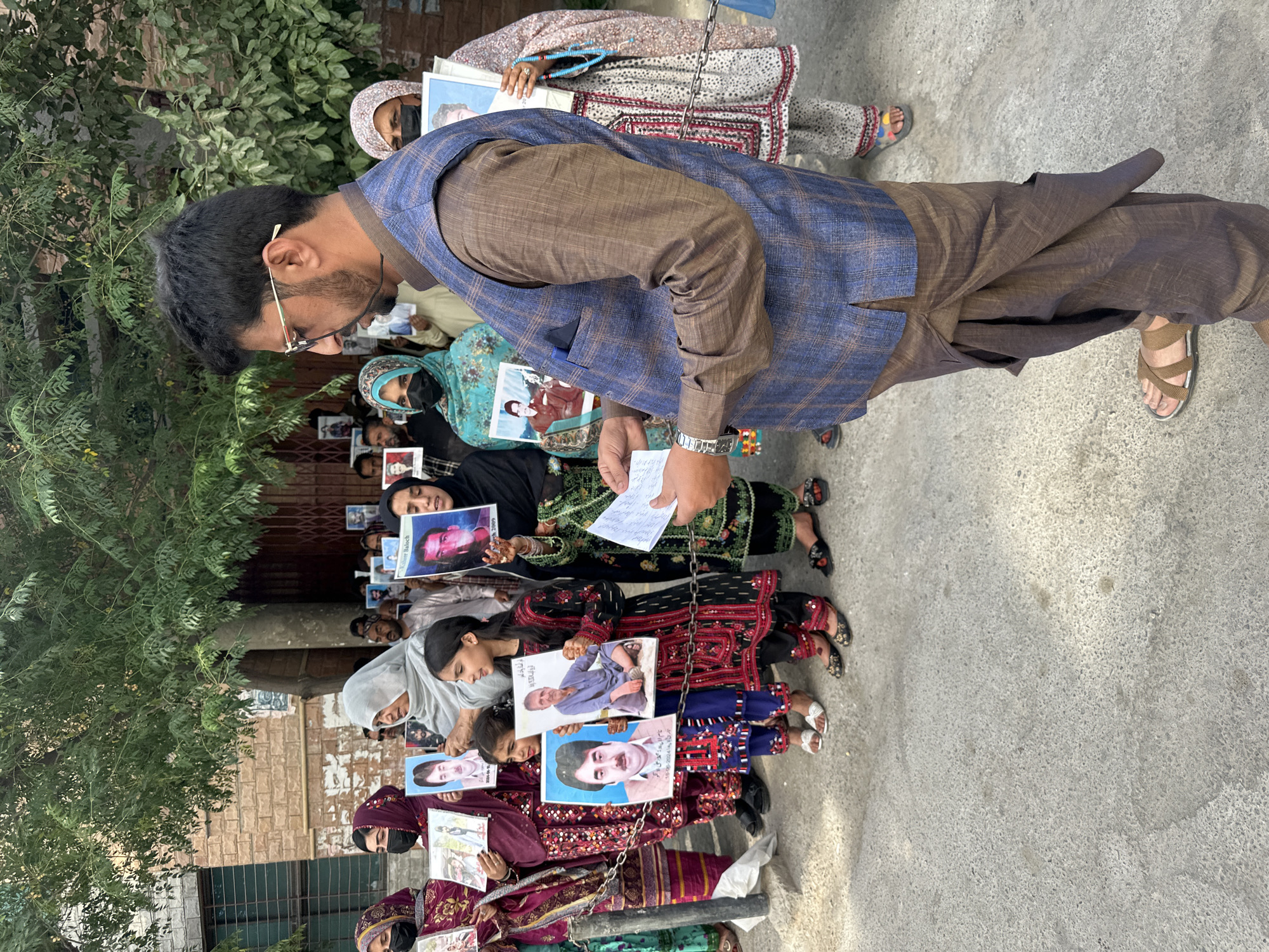 In Quetta City Tehsil, Pakistan, a group of people are demonstrating in what appears to be an outdoor urban setting. The demonstrators are holding up pictures of individuals. One man is visible at the front of the group, holding what appears to be a list or notes. The group is standing or sitting on a paved surface next to a building with green foliage visible in the background. Several demonstrators are wearing traditional clothing, and some are wearing face masks.
