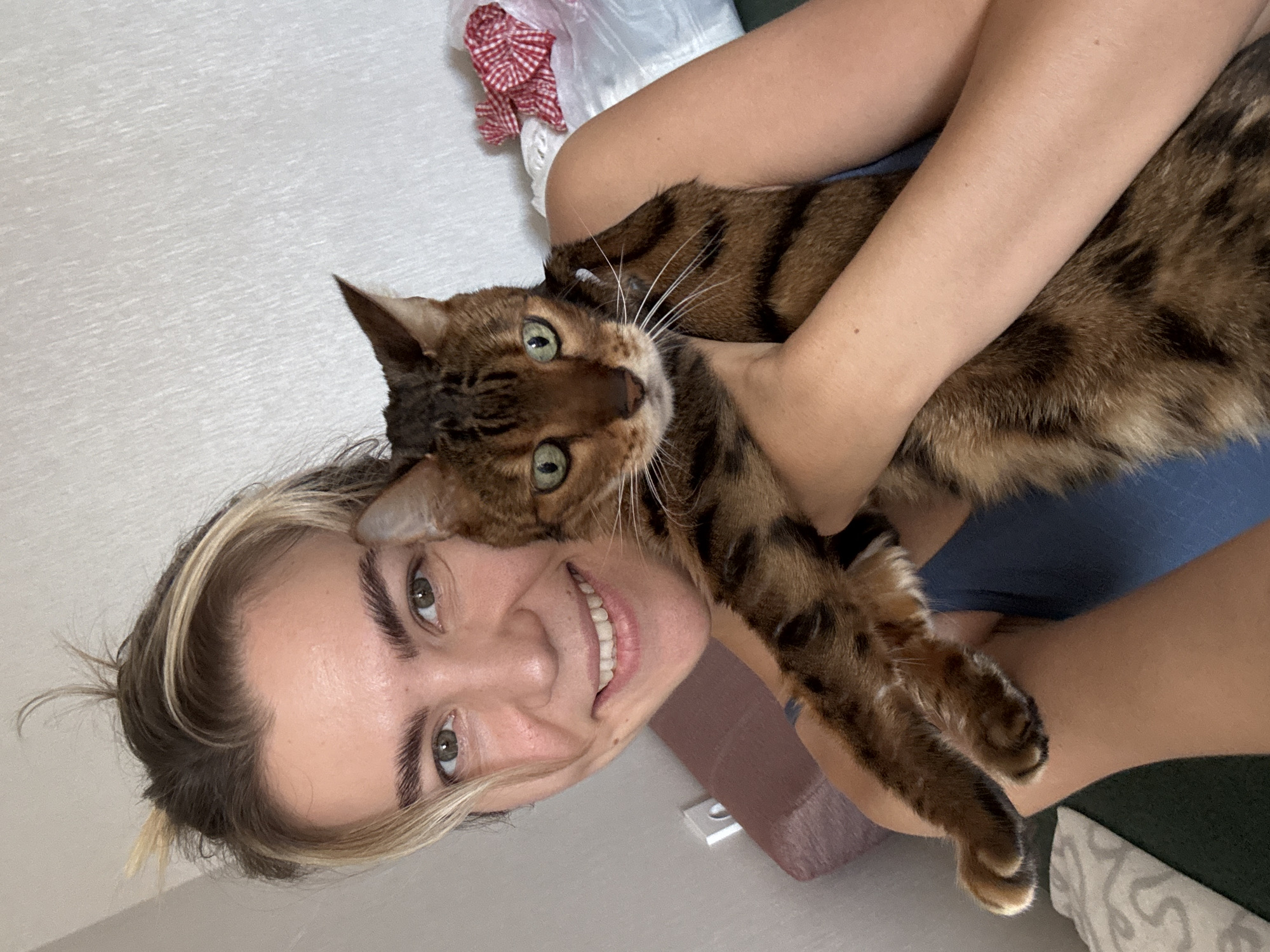 A young woman with short, blonde-streaked hair pulled back, is holding a Bengal cat in her arms and looking into the camera. She has green eyes, a wide smile, and is wearing a dark blue top. The cat is brown with black spots and stripes, has green eyes, and is looking directly at the camera. The background appears to be an indoor setting with a textured white wall and a glimpse of furniture. The lighting is natural and the overall impression is a candid selfie.
