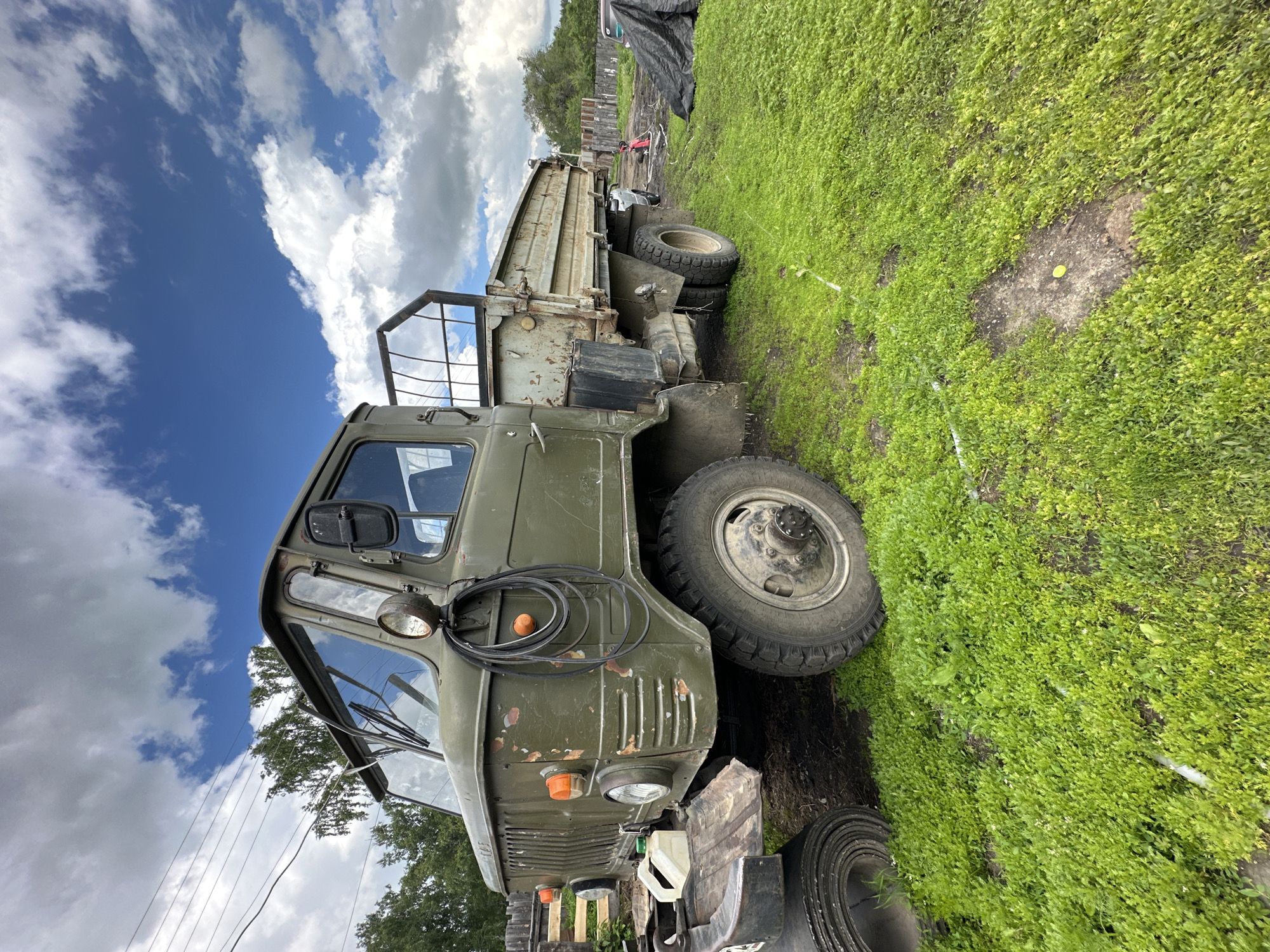 An olive-green, four-door truck is parked on a patch of green grass under a cloudy blue sky. The truck is older, with visible rust on its exterior, particularly on the front grill and along the sides of the bed. The truck has large, sturdy tires, with dual wheels on the rear axle. The bed of the truck is empty, with a metal grate at the front. The cab has two side mirrors, a single headlight on the passenger side, and two orange turn signals on the front grill. A coiled black hose is hanging from the side of the cab. In the background, there are trees, a wooden fence, and a black tarp covering something on the ground. The truck appears to be stationary and not in use.