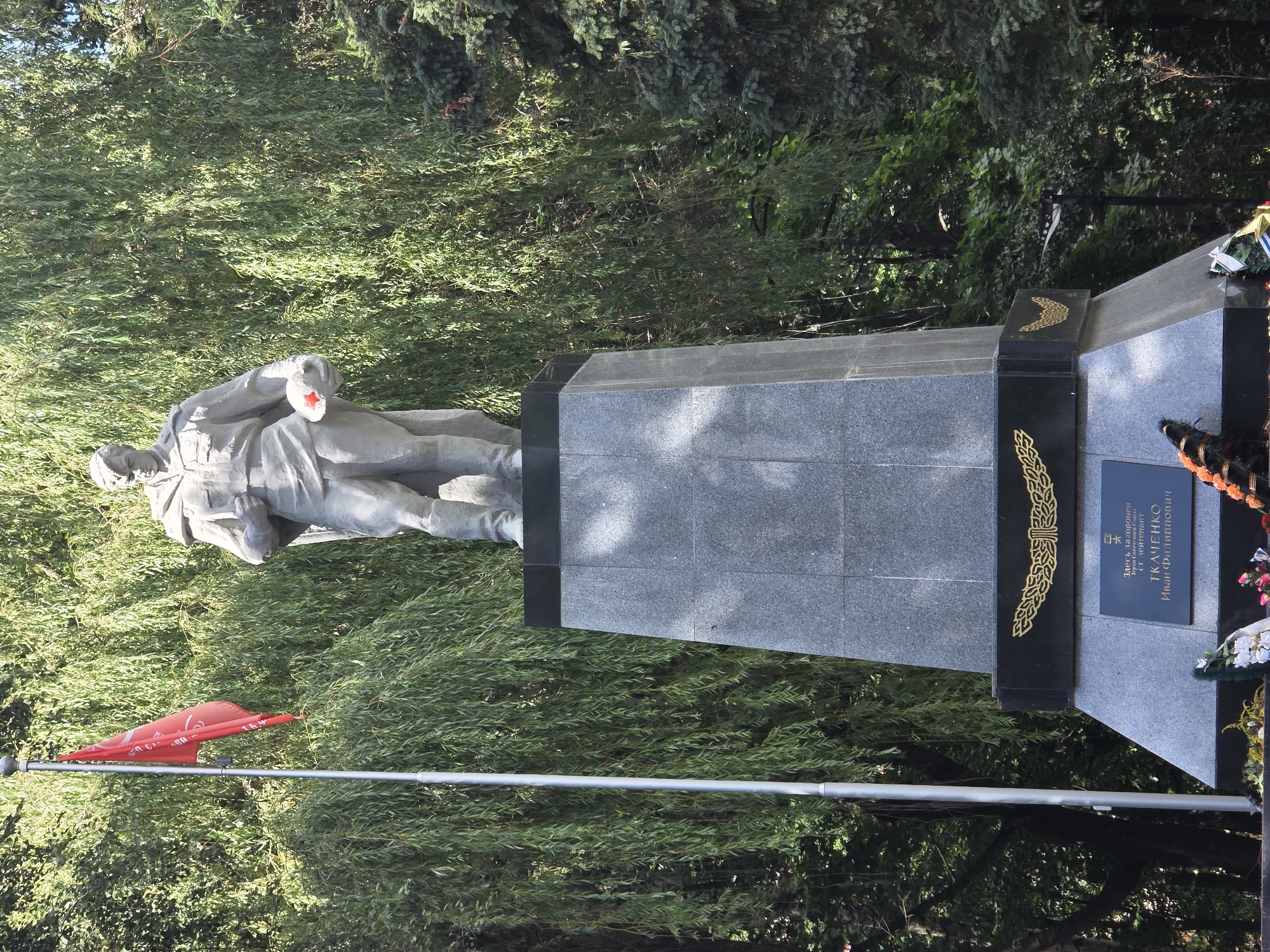 The image shows a monument to Ivan Filippovich Tkachenko, a Hero of the Soviet Union, located in Zelenogradsk, Russia. The monument features a gray stone statue of a soldier in uniform holding a helmet with a red star. The statue stands on a large, stepped granite pedestal. In front of the pedestal, there are wreaths of flowers, some of which appear to be artificial. To the left of the monument, a red flag flies on a tall pole. The background consists of lush green trees, suggesting an outdoor, possibly park-like setting. The lighting indicates it is daytime.