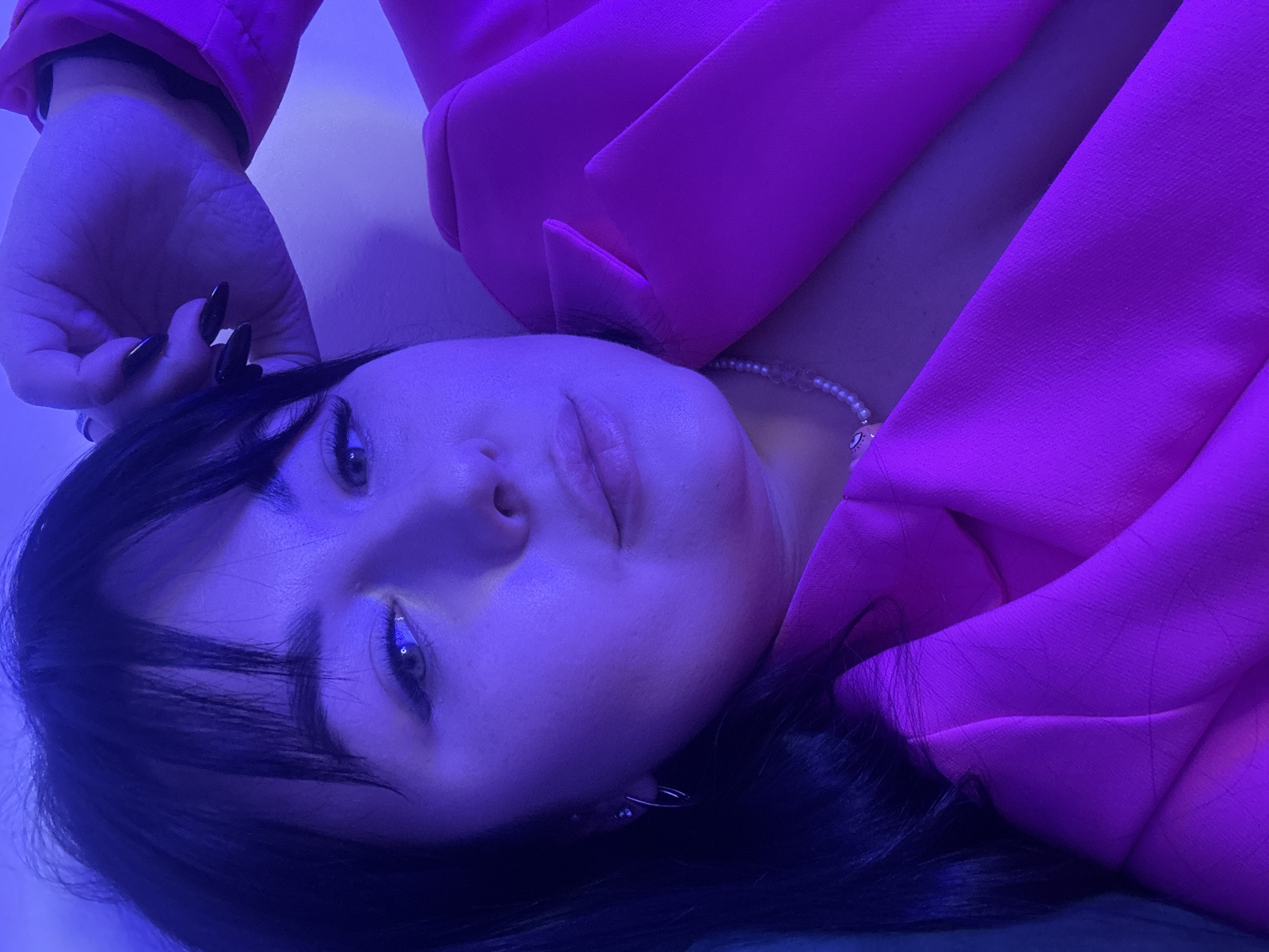 A woman with dark hair and bangs is lying down wearing a pink blazer. She is facing upwards with her left hand raised near her head. She is wearing a necklace. The lighting in the scene is predominantly blue and pink. The location is not discernible from the image.