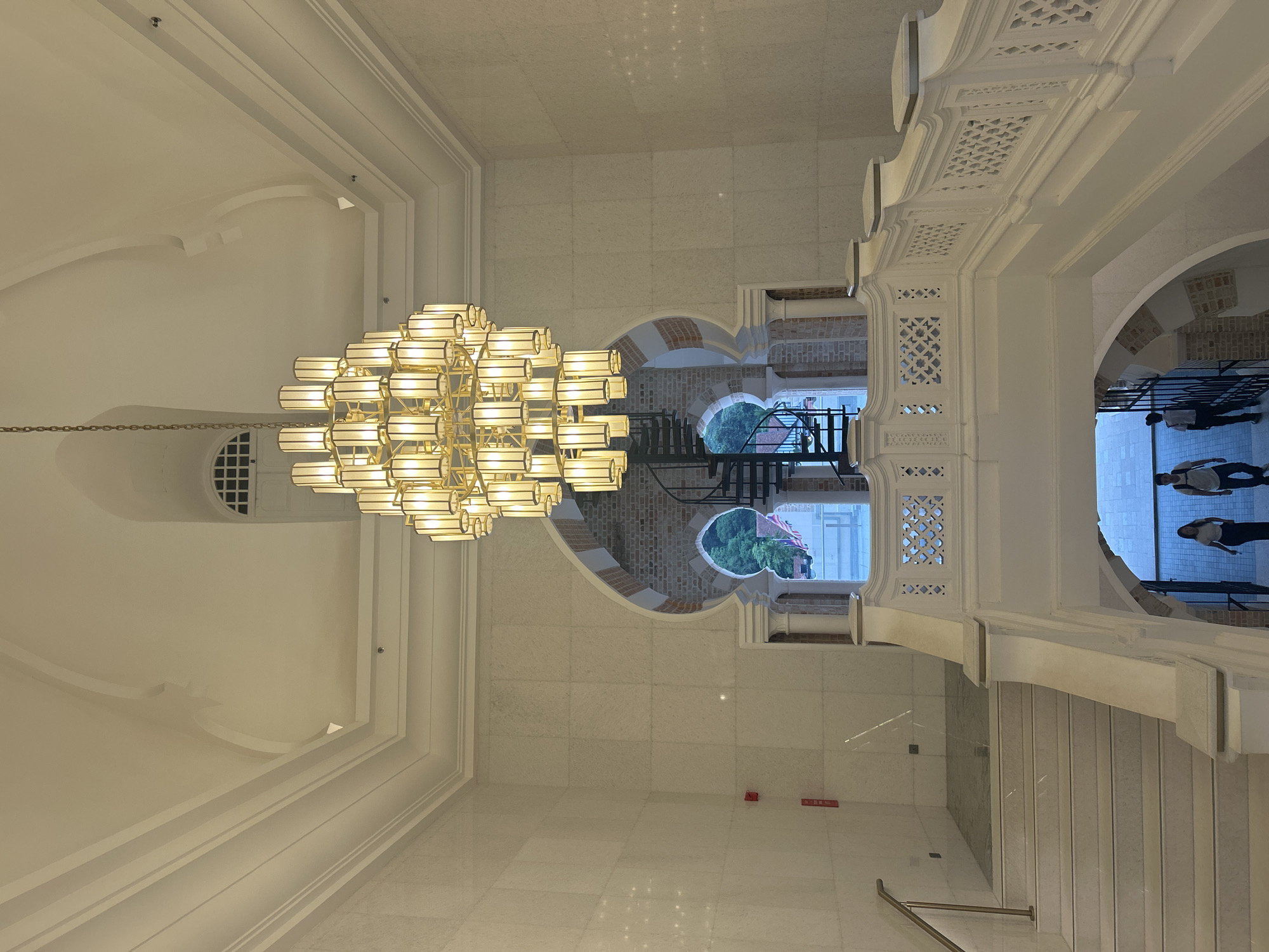 The image shows the interior of a grand staircase and atrium, likely in Kuala Lumpur, Malaysia, given the architectural style which evokes colonial-era Malaysian buildings. A large, multi-tiered chandelier hangs prominently in the center of the frame, illuminating the white marble walls and ornate white balustrade of the staircase. The staircase itself is wide and sweeps downwards, with a marble finish and decorative cutouts in the balustrade.
In the background, through an arched opening with Islamic architectural influences, a spiral staircase is visible, ascending towards a window overlooking lush green trees. The window above the arched opening features a grid pattern. At the bottom of the visible staircase, two individuals, a man and a woman, are walking away from the camera, one carrying a backpack. The walls are clad in large, light-colored marble tiles. A red fire alarm is visible on the left wall. The overall impression is one of opulence and historical grandeur within a public building.