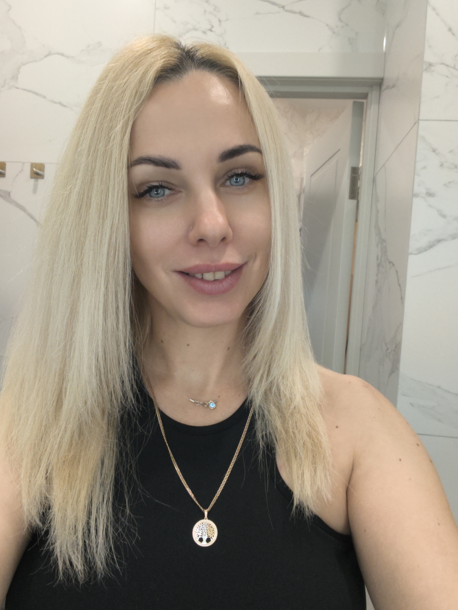 A woman with long, blonde hair and blue eyes is the central figure in a selfie. She wears a black tank top and two gold necklaces, one with a "tree of life" pendant and the other with a small, light blue stone. The background features a white marble wall and a partially visible white door. The location is likely a bathroom, but the exact city, Krasnodar, Russia, is not verifiable from the image alone.