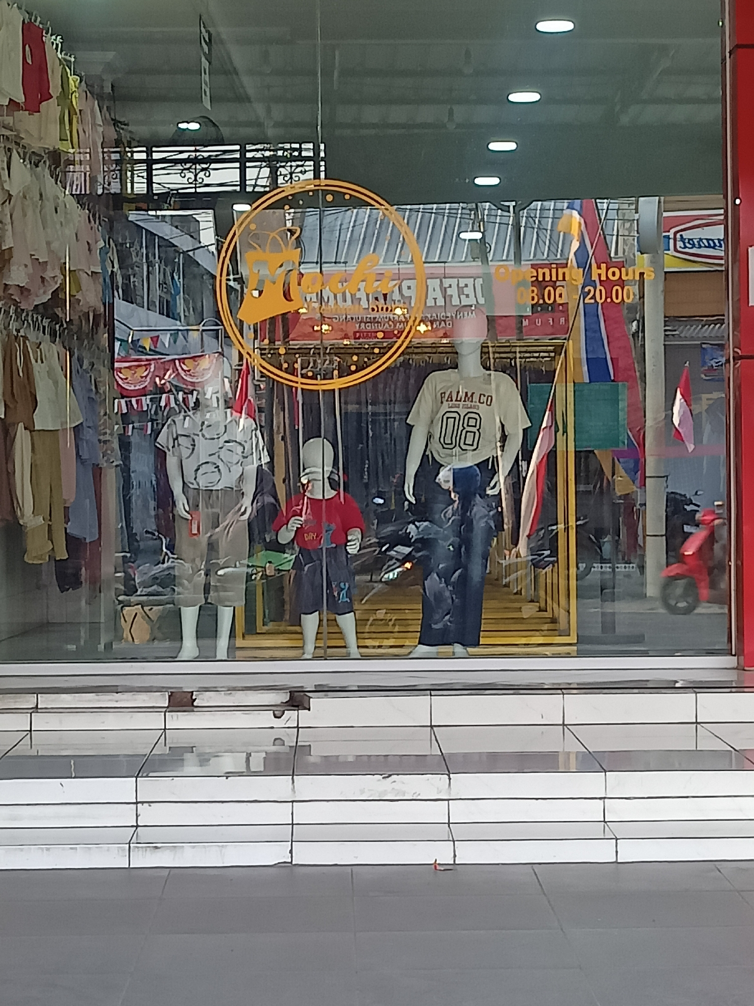 This image captures a street-level view of a storefront in Gemolong, Indonesia. The shop window displays mannequins dressed in children's and adult clothing. A circular logo with the text "Mocha" is visible on the window. Inside, racks of clothing are organized on display. The storefront is adorned with festive banners and flags. In the background, motorcycles are parked. The area in front of the store features a tiled walkway and steps leading up to the entrance. The overall scene suggests a retail environment in a commercial district.