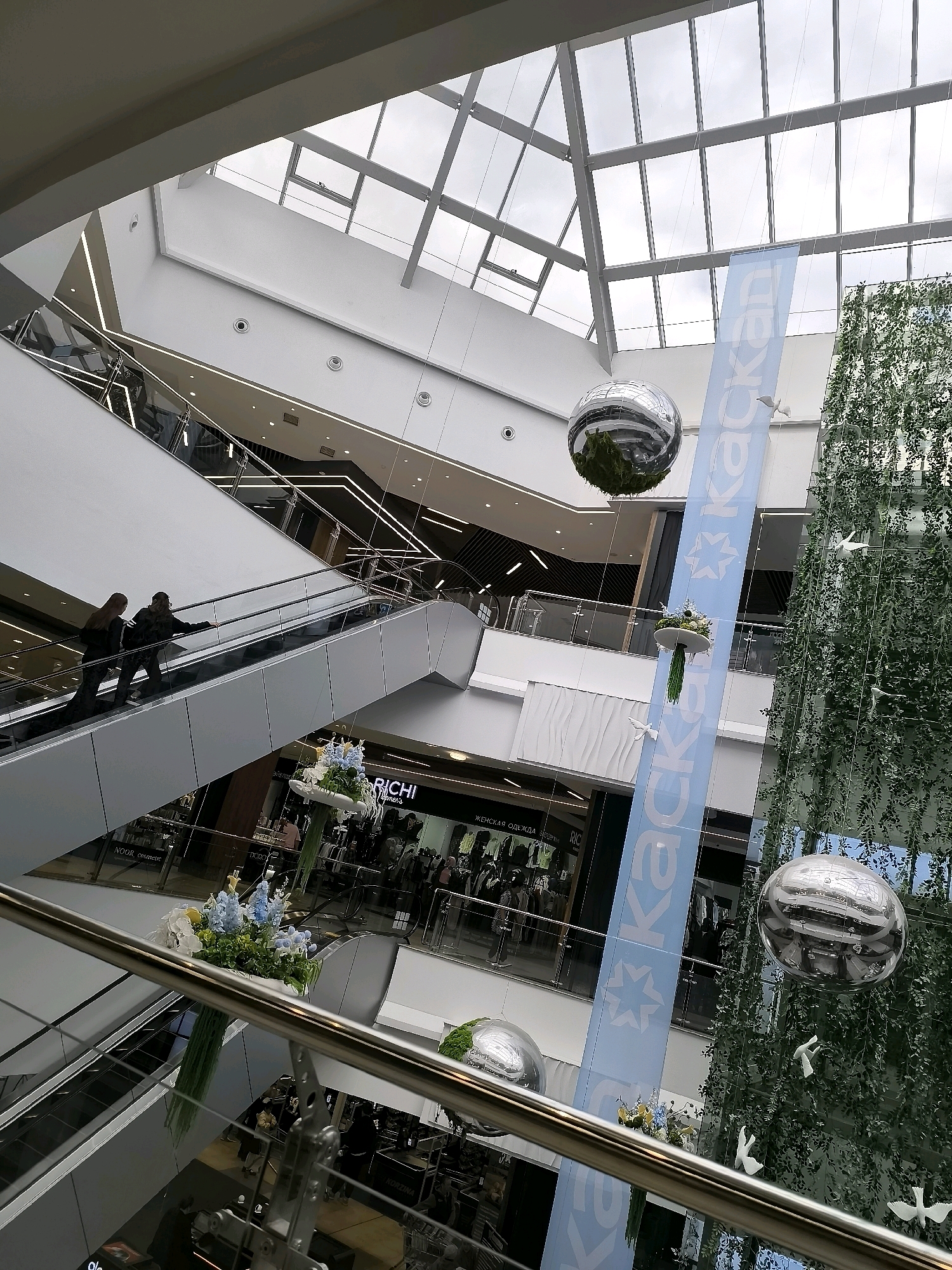 The image depicts the interior of a modern shopping mall, characterized by its multi-level design and contemporary architecture. A prominent feature is a large, light-blue banner hanging vertically, bearing white text and a star-like logo, likely for a retail establishment. Suspended from the ceiling or upper levels are several large, spherical mirrored decorations, one of which reflects the surrounding environment. A tall, vertical structure adorned with green foliage, resembling a living wall or a decorative tree, occupies a significant portion of the right side of the frame.
In the mid-ground, an escalator is visible, with two individuals ascending. They are positioned towards the left of the escalator. On the floors below, various shop fronts with glass facades are visible, displaying merchandise and signage. One sign clearly reads "RICHI Womens" and another "ЖЕНСКАЯ ОДЕЖДА" (Women's Clothing). Decorative floral arrangements, featuring blue and white flowers with green accents, are placed on ledges or balconies.
The overall environment is bright, with natural light filtering through a large, angled glass ceiling, revealing a cloudy sky. The mall's interior features clean lines, polished surfaces, and a sense of open space. The presence of escalators, shops, and decorative elements indicates a commercial and recreational setting. The image is taken from a slightly elevated perspective, looking down and across the mall's interior. No specific landmarks identifiable with Qarağandı city administration are visible.