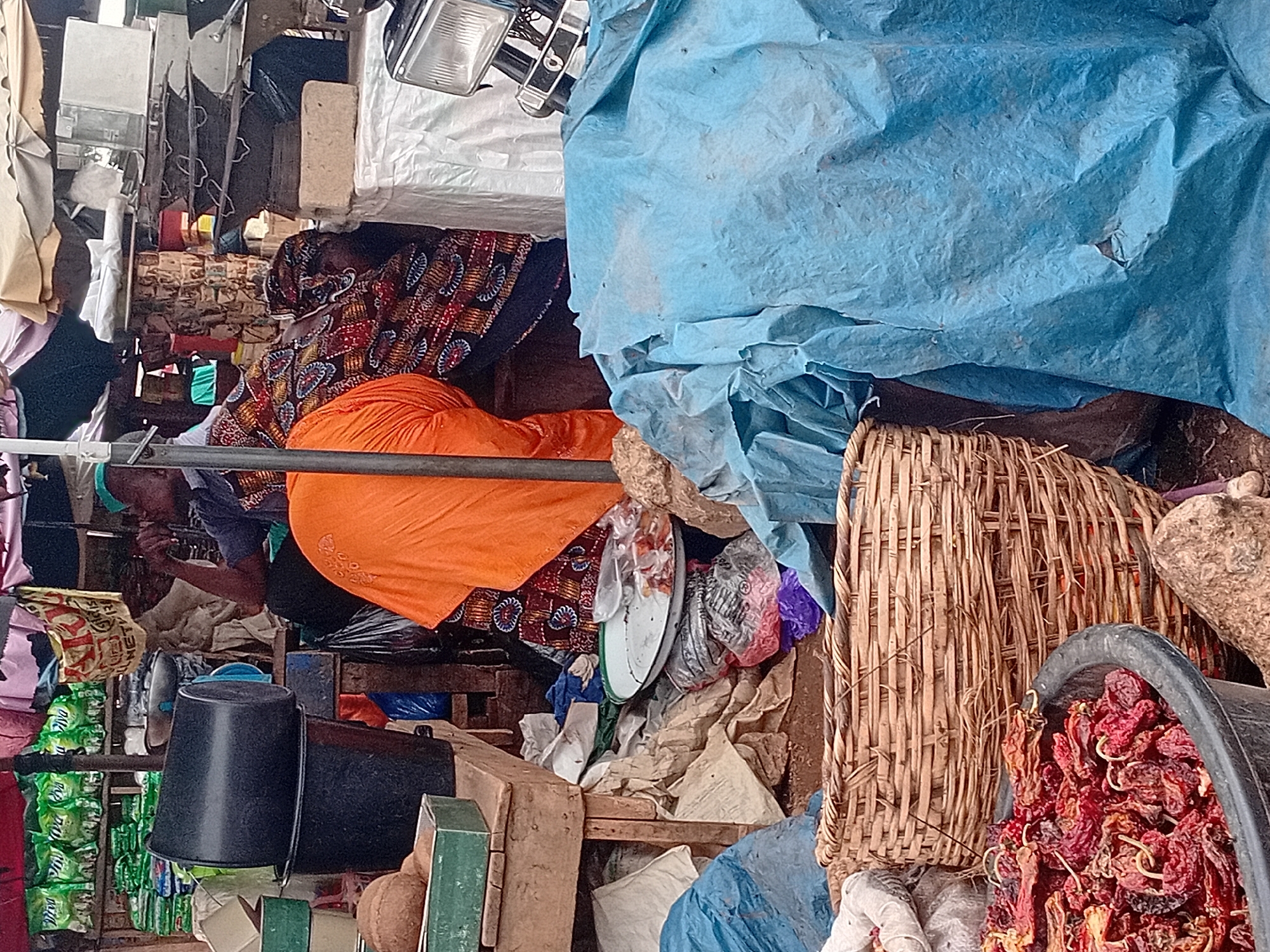 The image depicts an outdoor market stall in Maiburiji, Nigeria. In the foreground, a large, dark plastic basin is filled with dried red peppers. To the right of the basin is a woven basket, and behind it, a large sheet of blue tarpaulin is draped over various items. In the middle ground, a person wearing an orange garment and a patterned fabric headwrap is seated, partially obscured by the tarpaulin and other market goods. Another person, wearing a blue shirt and a green headcovering, is also seated and appears to be engaged in conversation or observing the surroundings. The background shows shelves stocked with green bags of what appears to be produce or packaged goods, along with various containers and market equipment. The overall scene suggests a bustling and typical marketplace environment.