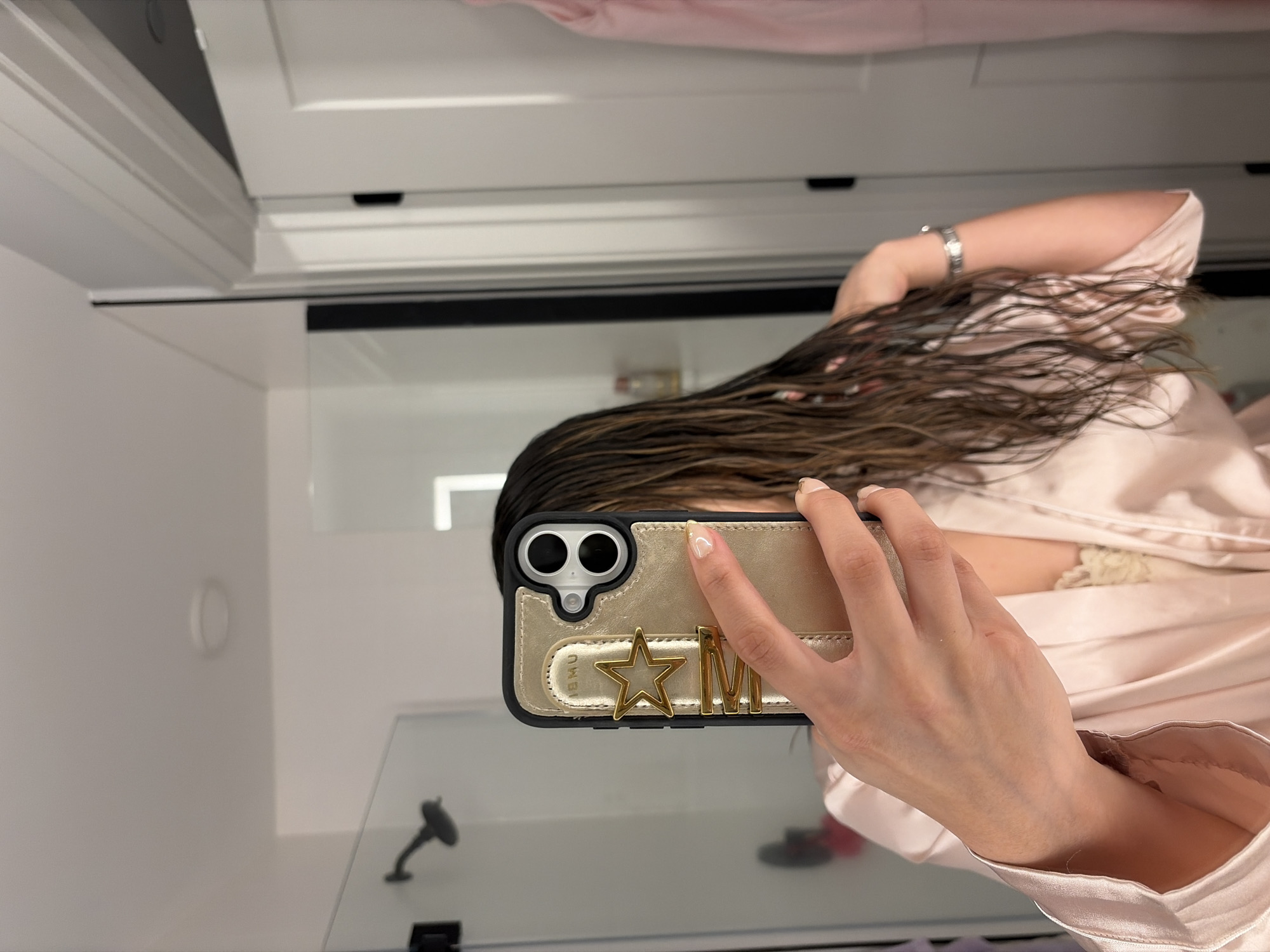 A person is depicted taking a selfie in a bathroom or dressing area in Austin, USA. The image is captured from an overhead perspective, with the person holding a smartphone to photograph their reflection in a mirror or reflective surface.
The subject's face is entirely obscured by the smartphone they are holding. Long, dark brown hair, appearing wet and somewhat messy, falls over their shoulders. The person is wearing a light pink, satin-like robe or nightgown, with a hint of a lace undergarment visible beneath it. A silver watch or bracelet is visible on their left wrist.
The smartphone is housed in a distinctive gold and black case. Adorning the back of the case are decorative gold charms: an open star shape and the capital letter "M". Small text, "JIEMU," is also visible on the gold section of the phone case. The hand holding the phone features neatly manicured nails with a light polish.
The background shows white walls and white cabinetry overhead. A black horizontal strip or ledge is present in the mid-ground. Reflections in the mirror or glass surface reveal a white circular object on the wall to the left, and a dark fixture, possibly a shower head or faucet, to the right. An L-shaped light fixture is also reflected above the phone. The wet appearance of the hair suggests the person may have recently showered or washed their hair.