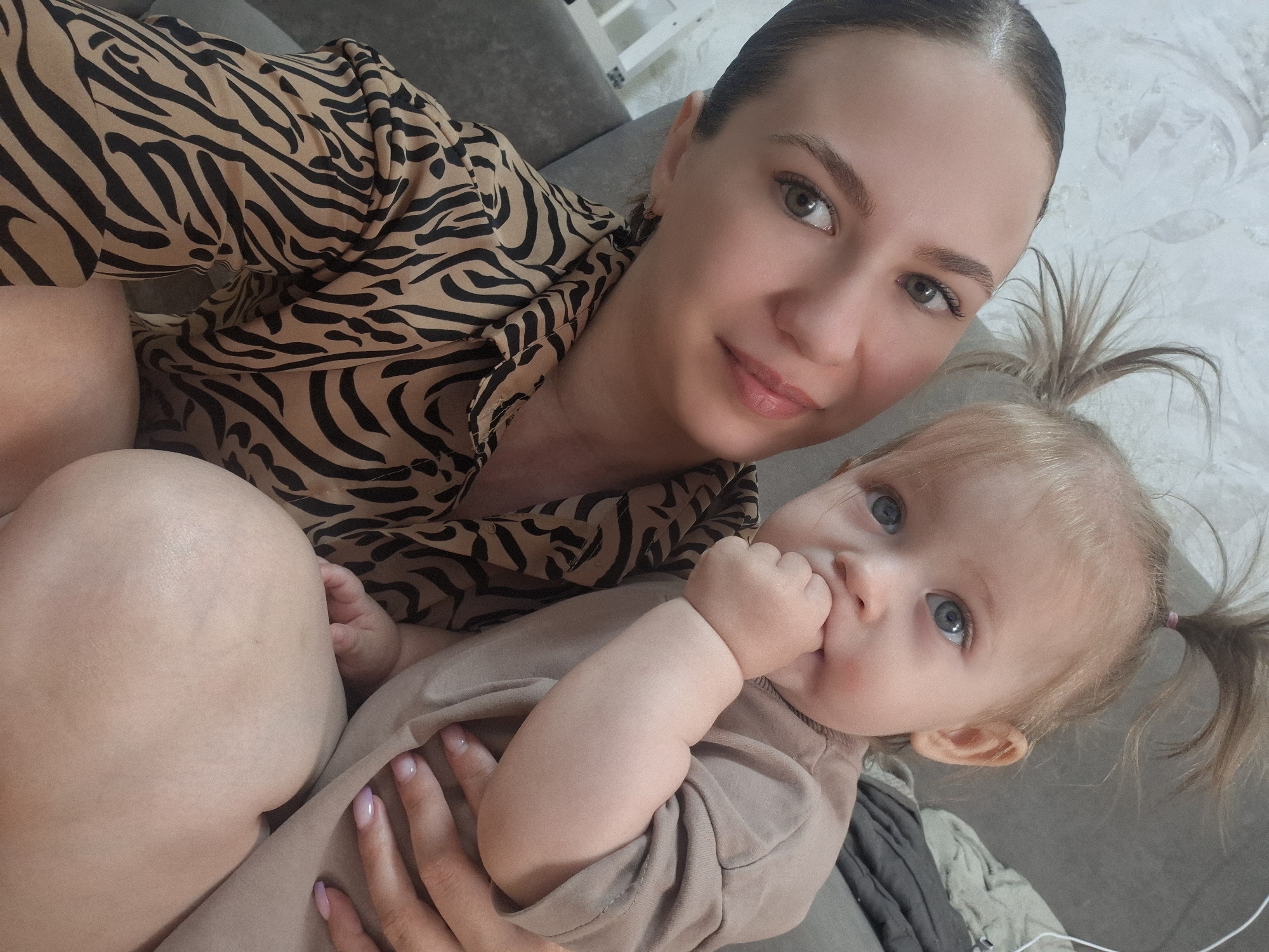 A selfie shows a fair-skinned woman with her hair pulled back, wearing a brown and black zebra-striped shirt, holding a fair-skinned baby with blonde hair styled in pigtails. The baby is wearing a beige onesie and has their hand in their mouth. The location appears to be indoors, with wallpaper visible behind the woman. The photograph may be taken in the city of Chaikovsky, Russia, but there are no identifiers in the image that confirm the location.