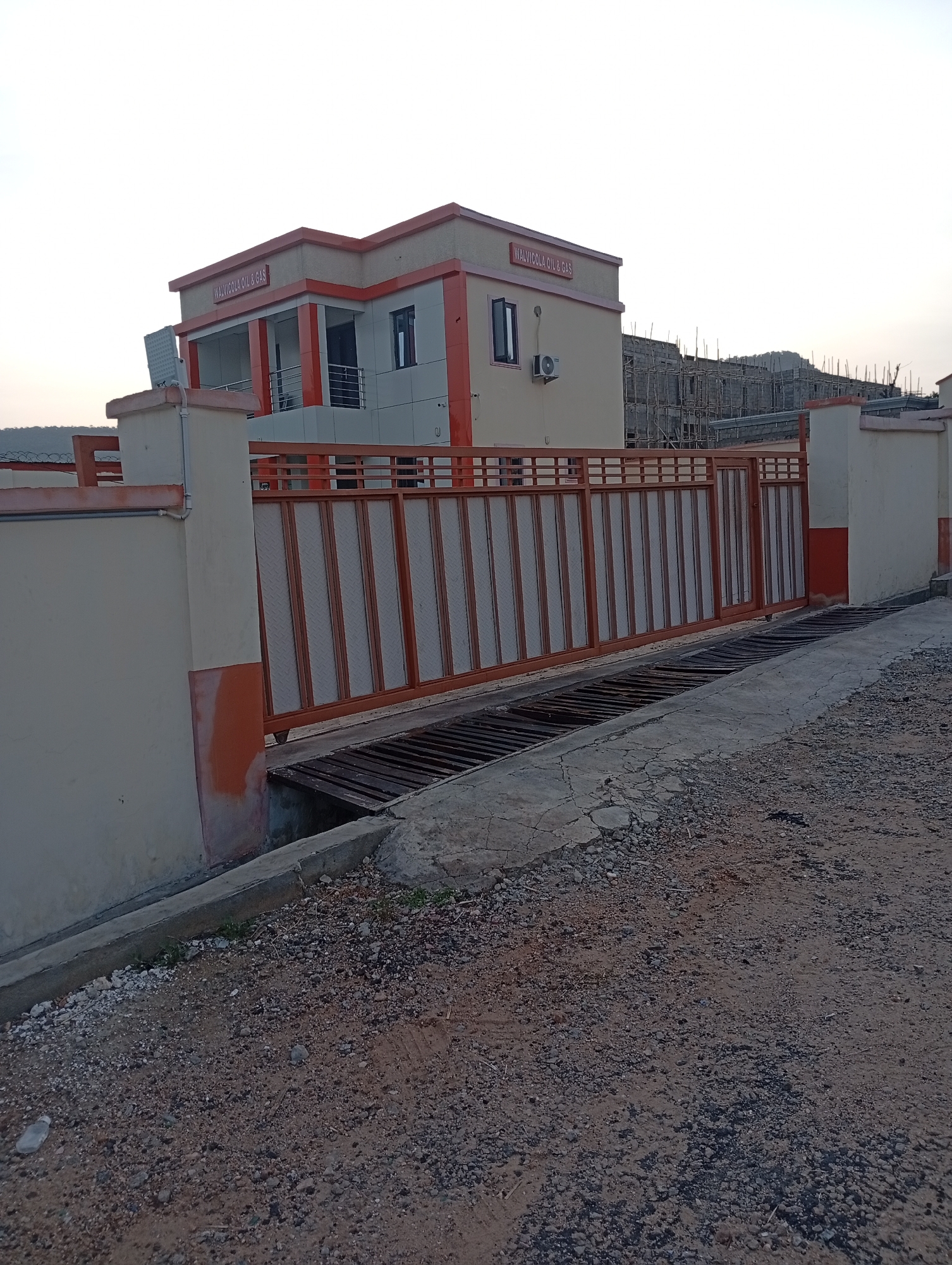 The image depicts the exterior of a two-story building with "WALVICOLA OIL & GAS" signage located in Adankolo, Nigeria. The building is painted in beige and orange, with a large sliding metal gate in front. The ground is unpaved and covered in gravel. A concrete drainage ditch runs along the side of the road. Construction scaffolding is visible in the background. No people are visible in the image.