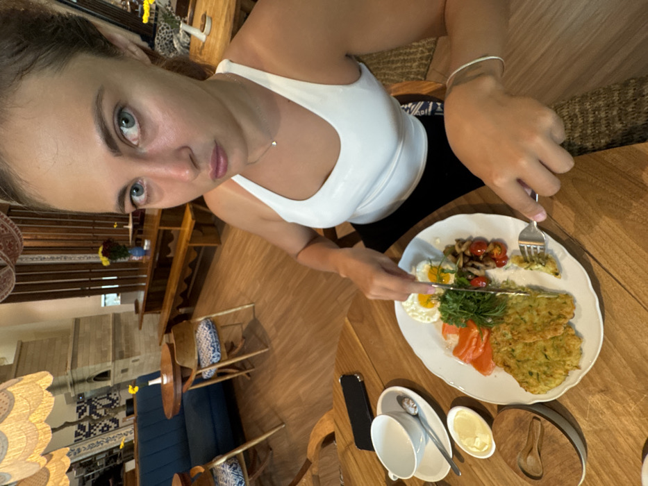 A young adult female, with light skin, dark hair pulled back, and wearing a white tank top with dark bottoms, is seated at a wooden table in an indoor restaurant or cafe in Kerobokan Kelod, Indonesia. She is looking directly towards the viewer with a neutral expression, her eyes appearing blue-green. A small silver heart-shaped pendant hangs from a chain around her neck, and a silver bracelet is visible on her left wrist.
Her hands are actively engaged in eating; she holds a silver fork in her left hand, which has a small piece of food on it, and a silver knife in her right hand, positioned above a white oval plate containing her meal. The meal consists of two fried eggs, several slices of salmon, a mound of fresh greens (likely rocket or similar) with dill, a portion of sautéed mushrooms and cherry tomatoes, and two flat, green-hued fritters.
To the left of her plate on the wooden table, a white teacup sits on a saucer with a spoon. Adjacent to this, a small white dish holds a pat of butter or spread. A dark-colored smartphone, possibly an iPhone, is placed face down on the table near the teacup. A small wooden container with a lid and a tiny wooden spoon rests closer to the edge of the table.
The background shows the interior of the establishment, characterized by wooden floors and a mix of seating arrangements. To the left, a blue upholstered sofa booth is paired with a round wooden table and two wooden chairs featuring patterned blue and white cushions. Further back, a long, light-colored wooden communal table with matching benches is visible. Decorative elements include a large, ornate light fixture with a scalloped, possibly woven, shade hanging from the ceiling. A light-colored, possibly brick or stone, structure is visible on the far left. Dark wooden paneling or screens adorn the walls, and small vases with yellow and red flowers are placed on tables throughout the space, contributing to a rustic yet refined ambiance.