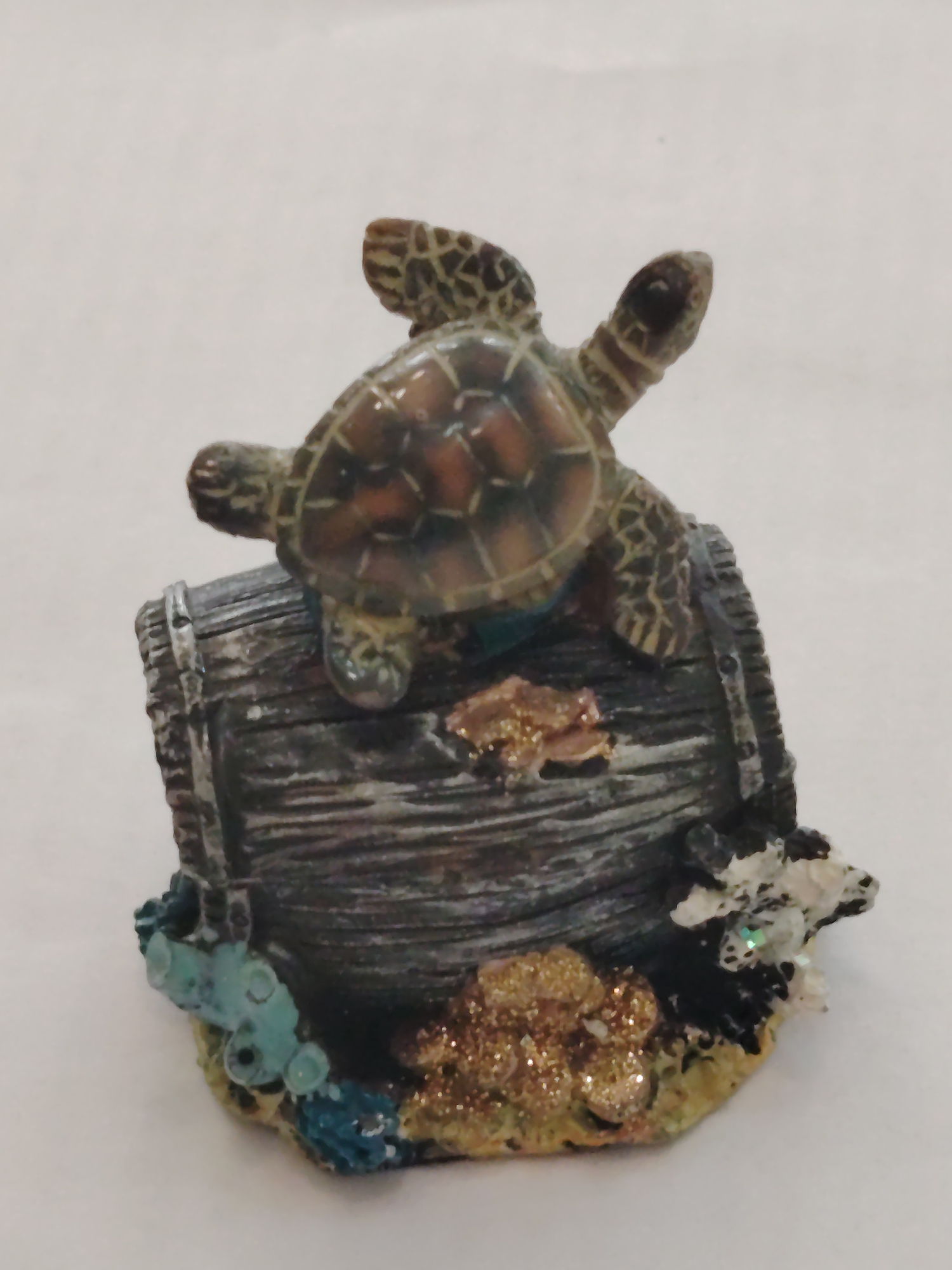 A turtle figurine rests on top of a miniature, weathered, gray barrel. At the base of the barrel are decorative coral and gold-colored glitter embellishments. The image appears to be taken against a plain, light-colored backdrop. No humans are visible. The location is not determinable from the visual content.