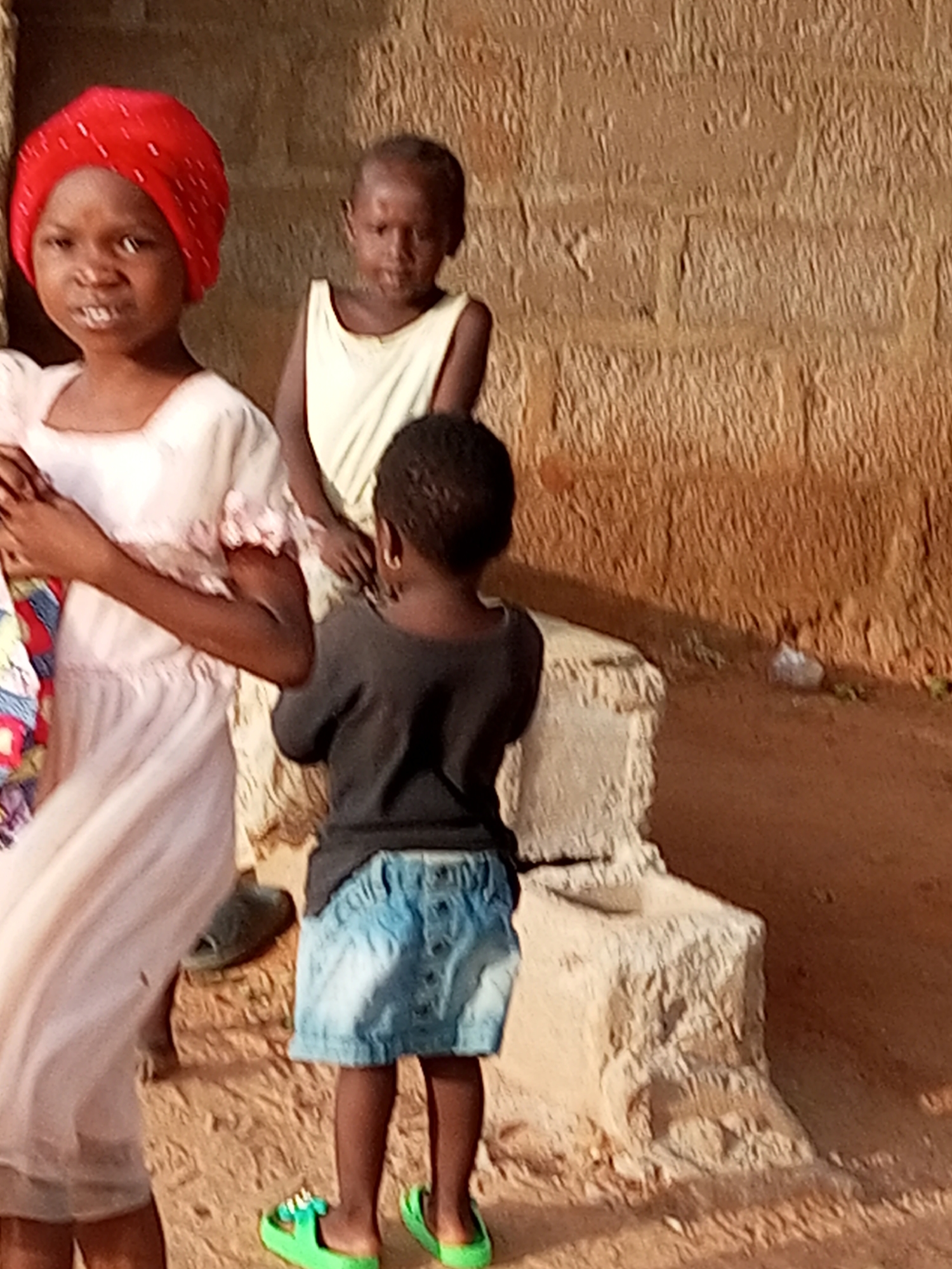 In a courtyard setting in Tempure, Nigeria, three young children stand near a textured, earthen wall. A girl in the foreground, wearing a bright red headscarf and a light-colored dress with pink floral accents, faces forward with a slight frown. To her right, another child in a pale yellow sleeveless top looks down. A third child, seen from behind, is wearing a dark t-shirt and a light blue denim skirt, with bright green sandals on their feet. The ground is sandy and reddish-brown, with a rough, light-colored stone structure partially visible. A piece of white plastic bag lies on the ground near the wall.