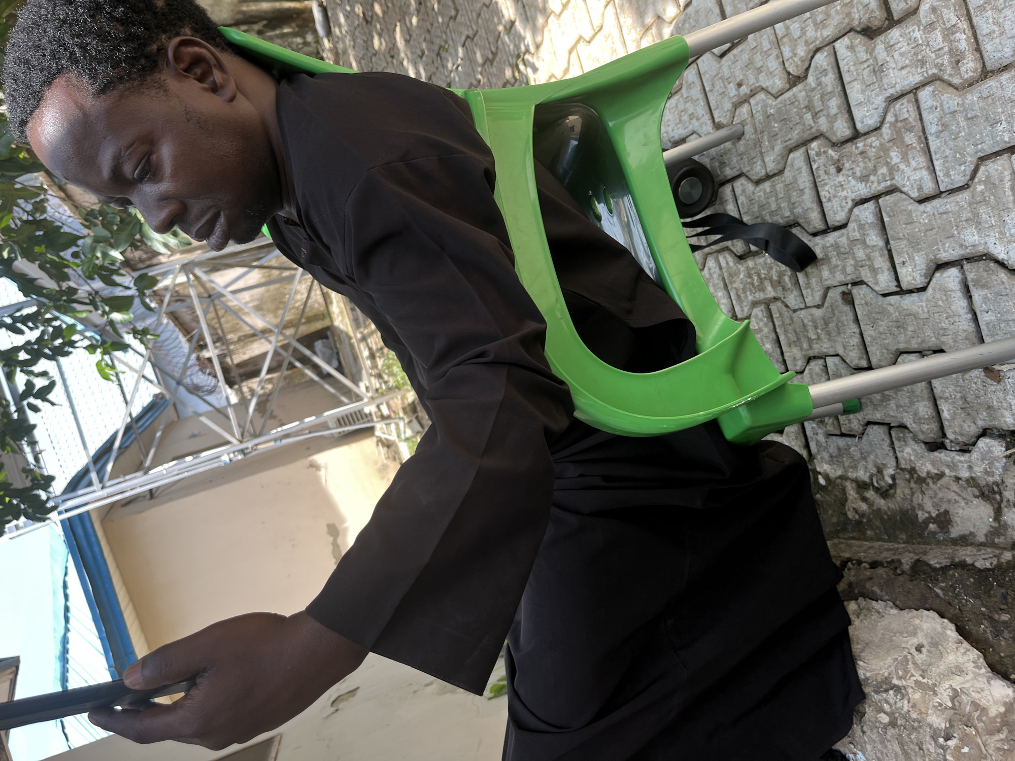 A man in black clothing sits on a green plastic chair outdoors in Galadimawa Village, Nigeria. He holds a black smartphone in his left hand and looks down at it. The man has dark skin and short, curly black hair. The chair is positioned on a paved area with interlocking grey pavers. In the background, a light-colored building with a blue roof is visible, along with metal scaffolding. Green foliage from trees hangs from the top of the frame.