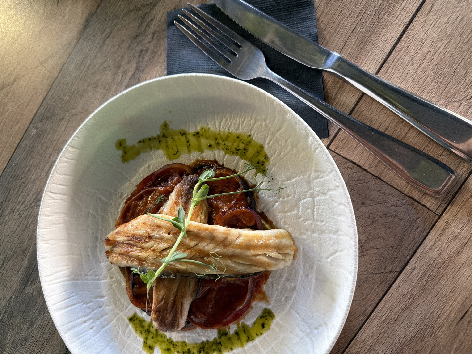 A top-down shot depicts a plate of food on a wooden table, with a fork and knife atop a dark gray napkin to the upper right of the plate. The plate is white with a slightly textured surface and a pale greenish tint. The food consists of what appears to be braised vegetables in a reddish sauce, topped with a piece of grilled fish and garnished with green sprigs. The sauce is also drizzled around the edge of the plate. The table has a wood-grain pattern running diagonally. The lighting is natural.