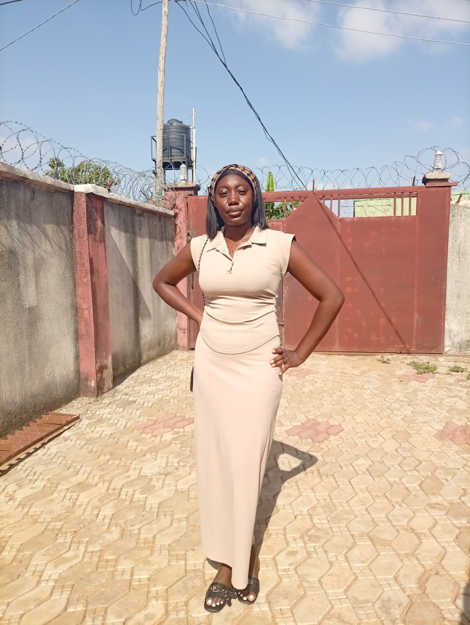A full-length shot depicts a woman standing outdoors. She wears a light brown dress and matching headband. Her hands rest on her hips. Behind her is a low wall topped with barbed wire. A gate is visible to the right of the wall and a water tank is in the background. The ground is paved with interlocking stone tiles. The location is Unknown, Unknown.