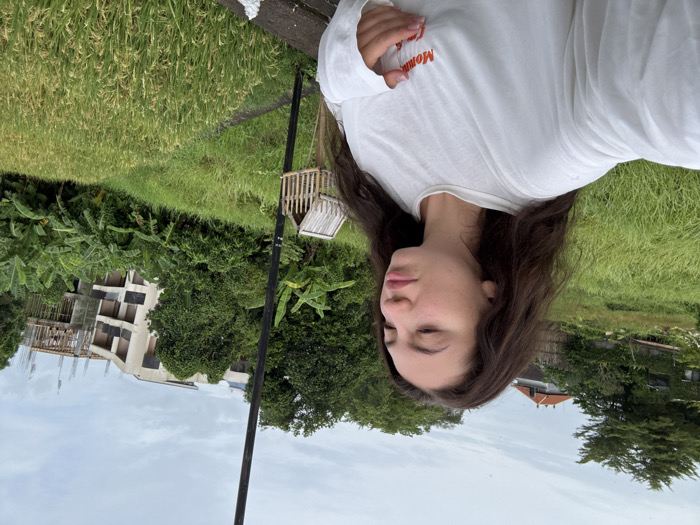 A young adult female with long, wavy dark brown hair and fair skin is depicted from the upper torso upwards, positioned slightly right of the frame's center. She wears a loose-fitting white long-sleeved shirt featuring faint red text, possibly "Mommy," on the left chest area. Her right hand is visible, partially obscured, near the shirt's front. She maintains a neutral expression, with her mouth slightly parted, gazing towards the right.
The scene is set outdoors in Dalung, Indonesia, under an overcast sky. The immediate foreground shows a dark, textured surface, possibly a concrete ledge. Below this, extensive vibrant green rice paddies dominate the lower right and mid-ground. A thin black pole stands vertically towards the center-right, bearing a small, light-colored wooden structure resembling a miniature house or offering box. Beyond the paddies and to the left, a section of wilder green grass and dense tropical vegetation is visible. The background reveals a mix of lush green trees and banana plants. Further back, two structures are discernible: a building with a reddish-orange tiled roof on the far left, partially obscured by trees, and a multi-story building under construction with exposed concrete framework and rebar towards the right side of the frame.