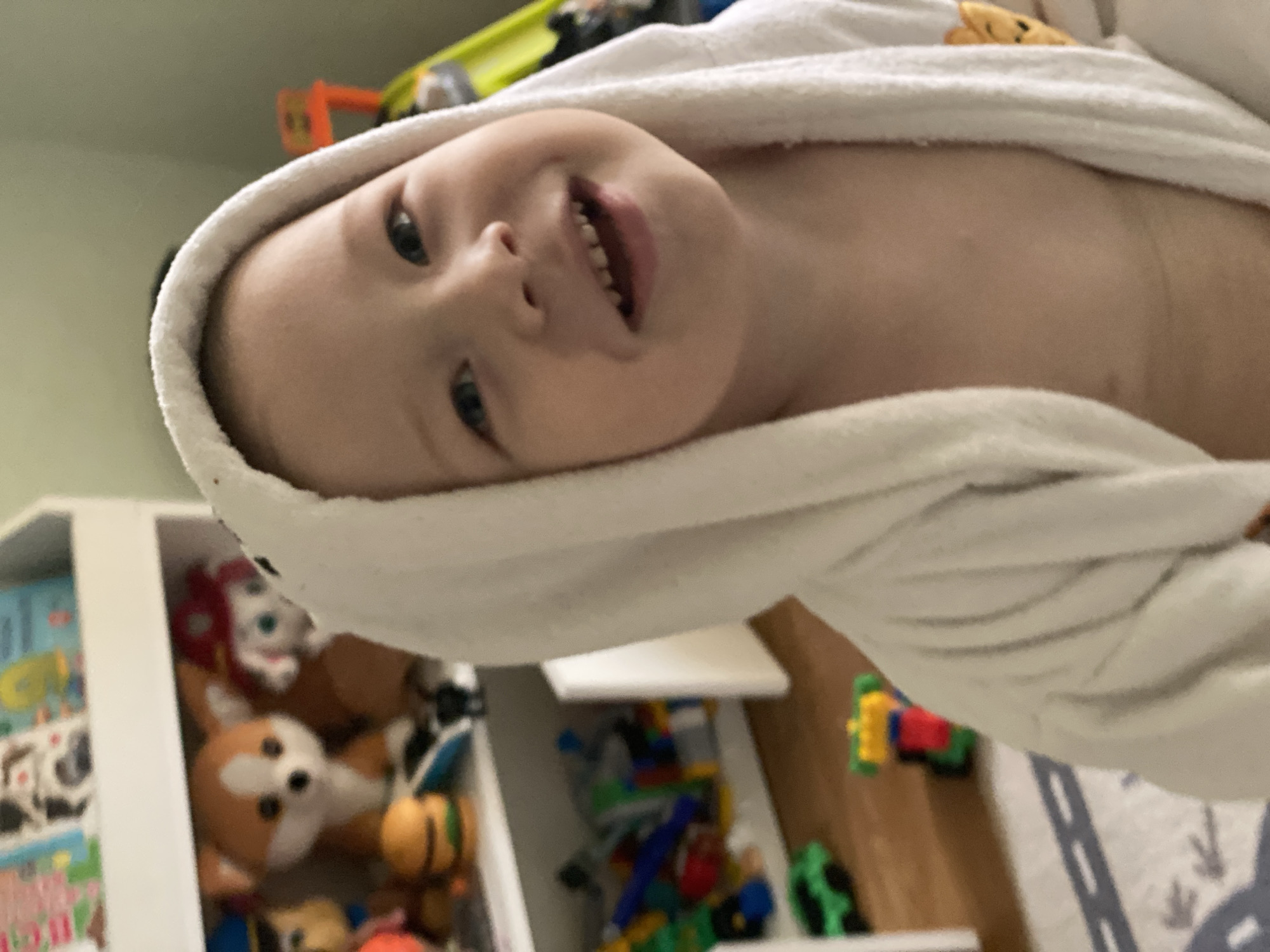 A young child is wrapped in a white towel, possibly after a bath, and is smiling at the camera. The child has fair skin, blue eyes, and light-colored hair. The background shows a child's room with shelves filled with toys and books. Some of the visible toys include a plush fox, building blocks, and toy cars. The photograph is slightly angled. There is no verifiable evidence indicating this photo was taken in Sochi, Russia.