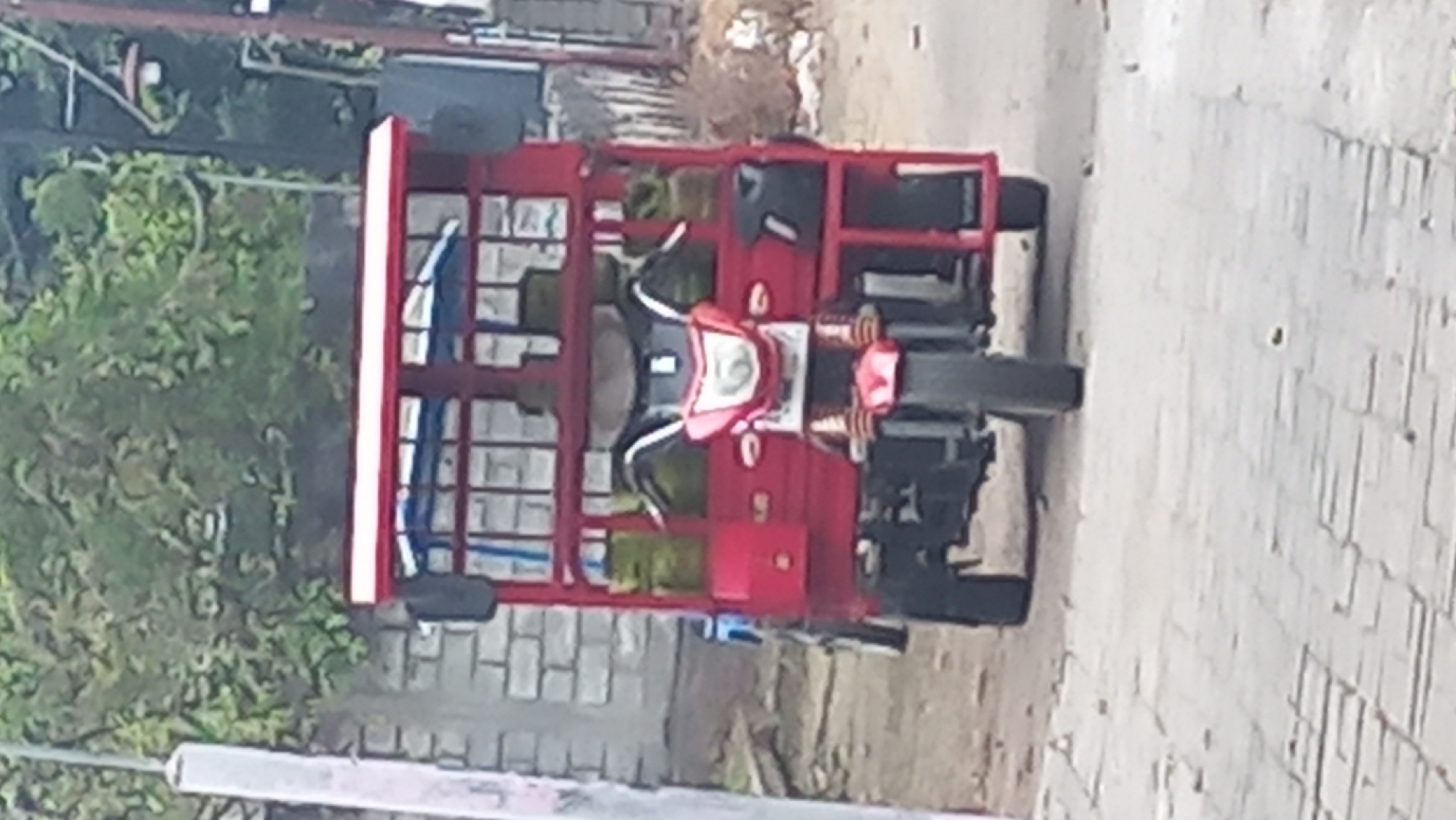 A three-wheeled red motorized vehicle, possibly a tuk-tuk or auto-rickshaw, is parked on a paved walkway. The vehicle has a covered passenger or cargo area with a red frame and a white roof. In the front, it has a motorcycle-like front end with a headlight and handlebars. The image appears to be taken from a street in Pare, Indonesia, with a brick wall and some foliage visible in the background.