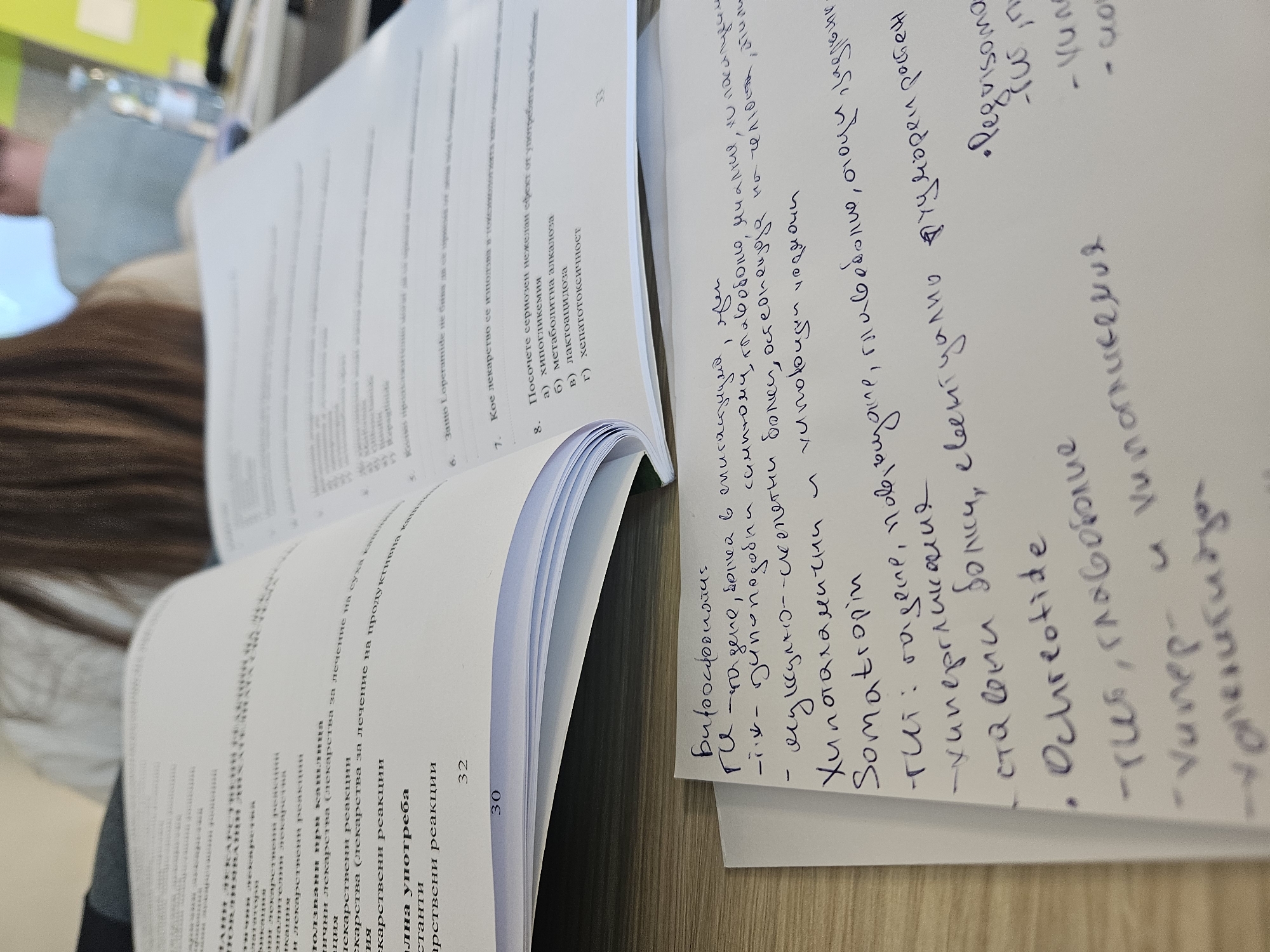 The image shows an open textbook in Bulgarian and a handwritten note lying on a table, likely in an academic or educational setting. The textbook is opened to a page discussing pharmaceutical reactions, and the note contains handwritten points about medications and their side effects. The angle and depth of field are shallow, suggesting the photo was taken at close range. The background is blurry but indicates the presence of another person. There is no clear indication that the location is in Varna, Bulgaria.