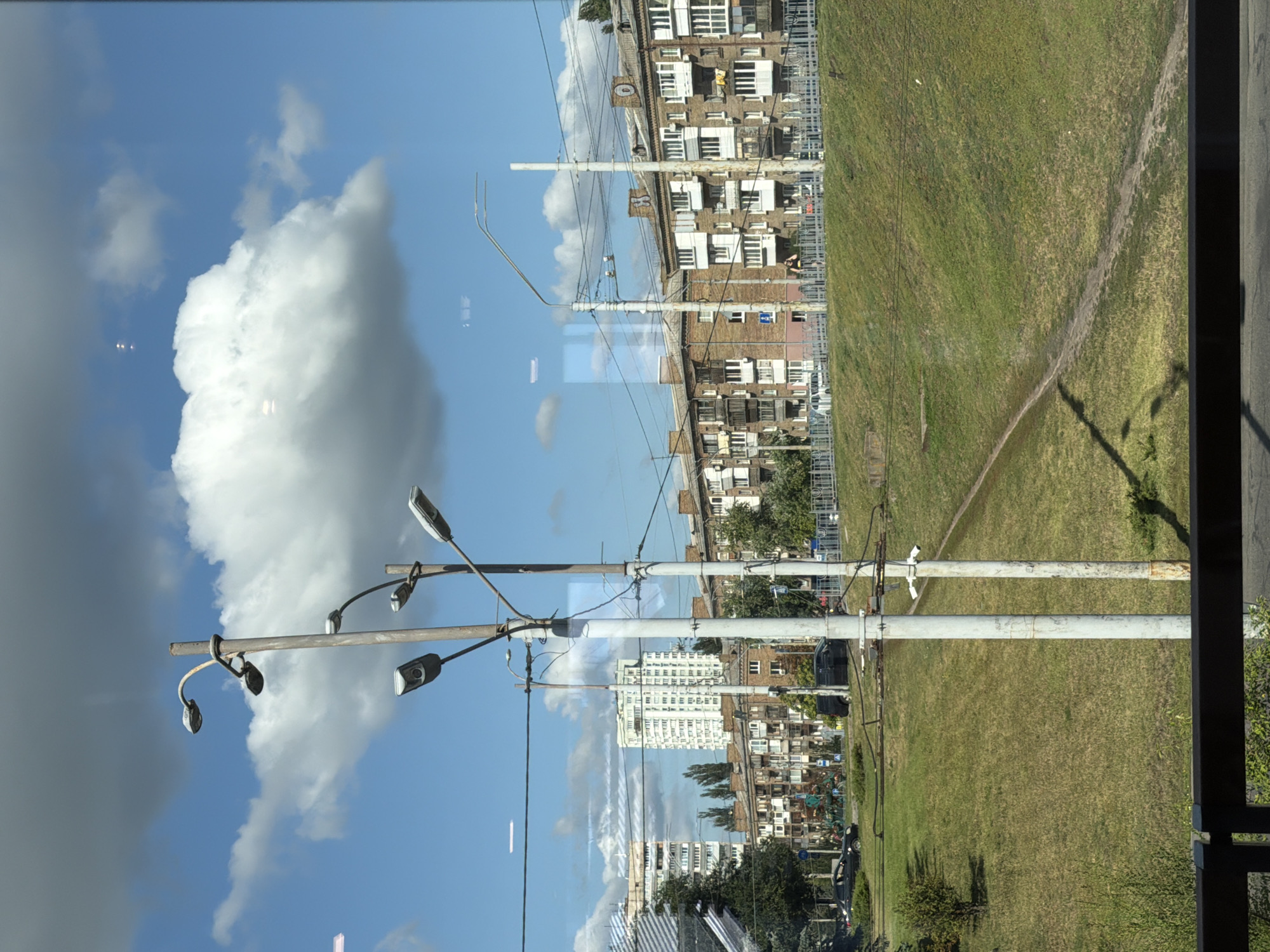 The image captures a view of a residential area in Kyiv, Ukraine, likely on a sunny day with scattered clouds. In the foreground, two tall, weathered lampposts stand prominently, one with multiple lights. Beyond them, a grassy slope leads up to a row of apartment buildings. These buildings appear to be of Soviet-era architecture, with brick facades and many balconies. A taller, modern white building stands further in the background. A paved path winds its way up the grassy slope. In the middle ground, there's a street with parked cars and some greenery. A dark metal railing is visible at the very bottom of the frame, suggesting the viewpoint is from an elevated position or a balcony. The sky is a clear blue, punctuated by fluffy white clouds.