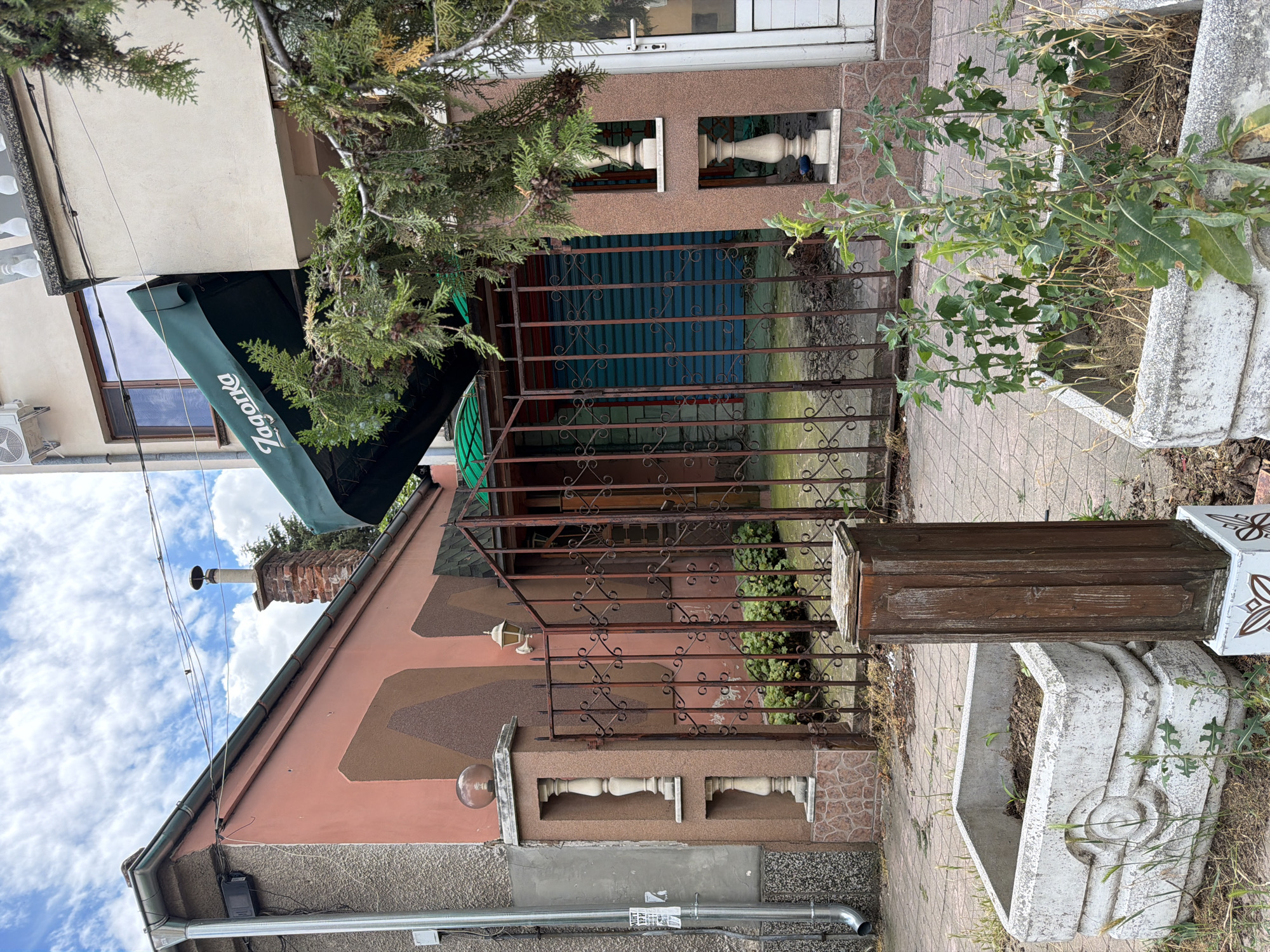 The image shows the exterior of a building with a green awning that reads "Zagorka". The awning is above a storefront with a blue metal roll-down gate. To the left of the storefront, there is a rusty metal gate leading to a small yard with some greenery. In the foreground, there are two concrete planters, one with some small plants and the other with dry grass and weeds. A wooden post stands between the planters. The building is made of light-colored plaster and has several decorative elements, including columns and ornate railings. The sky is cloudy. The image appears to have been taken in Pazardzhik, Bulgaria.