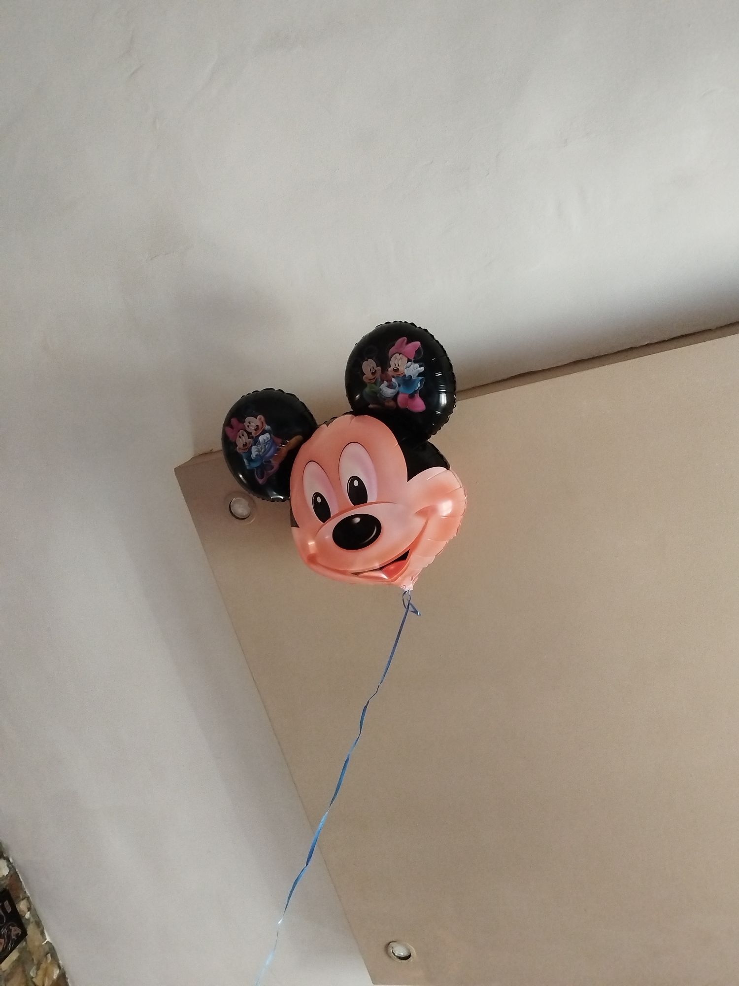 A Mickey Mouse balloon with two black balloons for ears floats below a ceiling in an indoor space. The balloon has a blue string. The location is Unknown, Unknown.