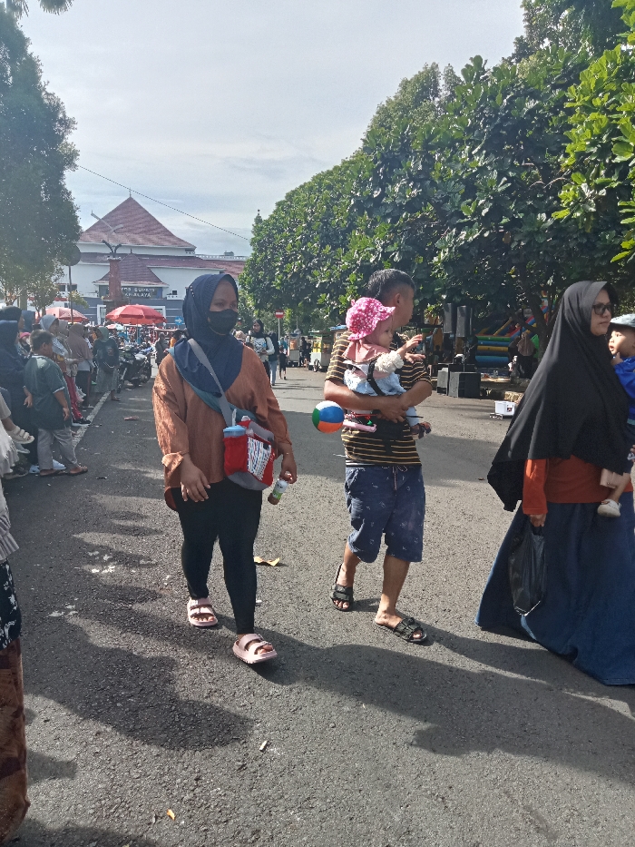 The image captures a bustling outdoor scene in what appears to be a public street or market area in Singaparna, Indonesia, under bright daylight. Strong shadows cast on the asphalt road suggest it is mid-morning to early afternoon.
In the foreground, three main groups of people are walking. On the left, a woman wearing a black hijab and face mask, an orange-brown long-sleeved shirt, black leggings, and pink sandals. She carries a red and white patterned sling bag across her chest and holds a small bottle in her right hand. To her right, a man with a short haircut, wearing a striped t-shirt, patterned blue shorts, and black sandals, carries a baby in a front-facing carrier. The baby wears a pink polka-dotted sun hat. The man also holds a colorful beach ball. Further to the right, partially visible, another woman in a black hijab, orange top, and long blue skirt walks, carrying a small child whose feet are visible near her skirt.
The background is crowded with numerous people. On the left, there's a dense gathering of individuals, with glimpses of stalls or vendors, including a red umbrella. In the distance, a large building with a distinctive red-tiled roof and white and blue walls is visible. Text on the building reads "BALAI SUK… AMALAYA," likely indicating "BALAI SUKAPURA." A line of lush green trees stretches along the right side of the road, providing shade. On the far right background, speakers and what appears to be a colorful inflatable bouncy castle or play area are discernible, suggesting recreational activities are part of the event. The ground shows some scattered litter, including cigarette butts and leaves.