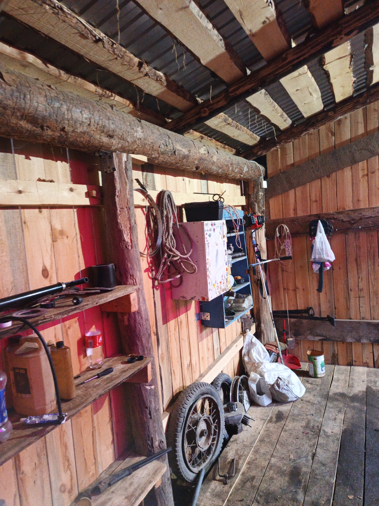 The image depicts the interior of a rustic wooden structure, likely a shed or garage, in Islambaevo, Russia. The space is cluttered with various tools and materials. A large, rough-hewn log spans across the ceiling, supported by wooden beams. The walls are constructed of vertical wooden planks, and the floor is made of weathered wooden boards.
On shelves mounted on the left wall, several items are visible, including a large brown plastic container, a smaller yellow plastic container, a glass bottle, a red and white can of Coca-Cola, a screwdriver, and a black cylindrical object. A long black pipe or rod lies horizontally on the top shelf.
To the right of the shelves, a blue shelving unit is mounted on the wall, holding various items. A large bundle of rope hangs from a hook on the wall above this unit. Further to the right, a white plastic bag containing items hangs on the wall, and below it, a black object, possibly a tool or part, is attached to the wall.
On the floor in the foreground and mid-ground, there are several items, including two wheels, one large motorcycle wheel with a tire and spokes prominently displayed. There are also several white plastic bags filled with materials, a red shovel, and a green and white cylindrical container. A long, thin dark object, possibly a rifle or a long tool, is placed on a wooden plank on the floor in the lower right section of the image. The lighting suggests it is daytime, with shadows cast by the objects and structures.