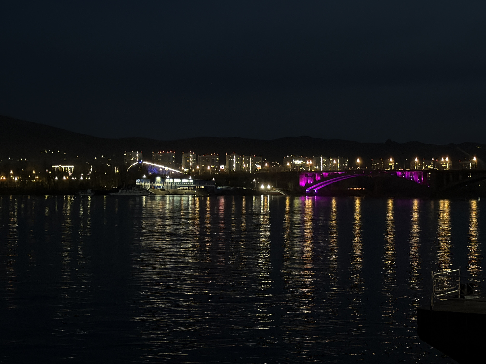 The image displays a night view of the city of Krasnoyarsk, Russia, seen across a wide body of water, likely the Yenisei River. The scene is dominated by the deep blue-black sky above and the reflective dark water below, with the illuminated city stretching across the distant bank.
On the right side of the distant shore, a prominent bridge is illuminated with striking purple and pink lights, casting vibrant, elongated reflections onto the water's surface. Along the rest of the city's skyline, numerous buildings feature illuminated windows and exterior lights, primarily in warm yellow and white tones. These city lights create a sparkling panorama against the dark backdrop of hills or mountains that form the horizon. Near the center-left of the distant bank, a larger structure, possibly a building or complex by the water, glows with brighter white and blue lights.
The foreground and midground are covered by the calm, dark water, which beautifully mirrors the city's illumination, showing shimmering vertical streaks of light distorted by gentle ripples. In the bottom right corner, a partial view of a dark structure, possibly a dock or a moored vessel, with a simple metal railing, is visible.
The image captures the urban landscape at night, devoid of people or specific activities, highlighting the serene beauty of the illuminated city reflected on the water. No visible text is present in the image.