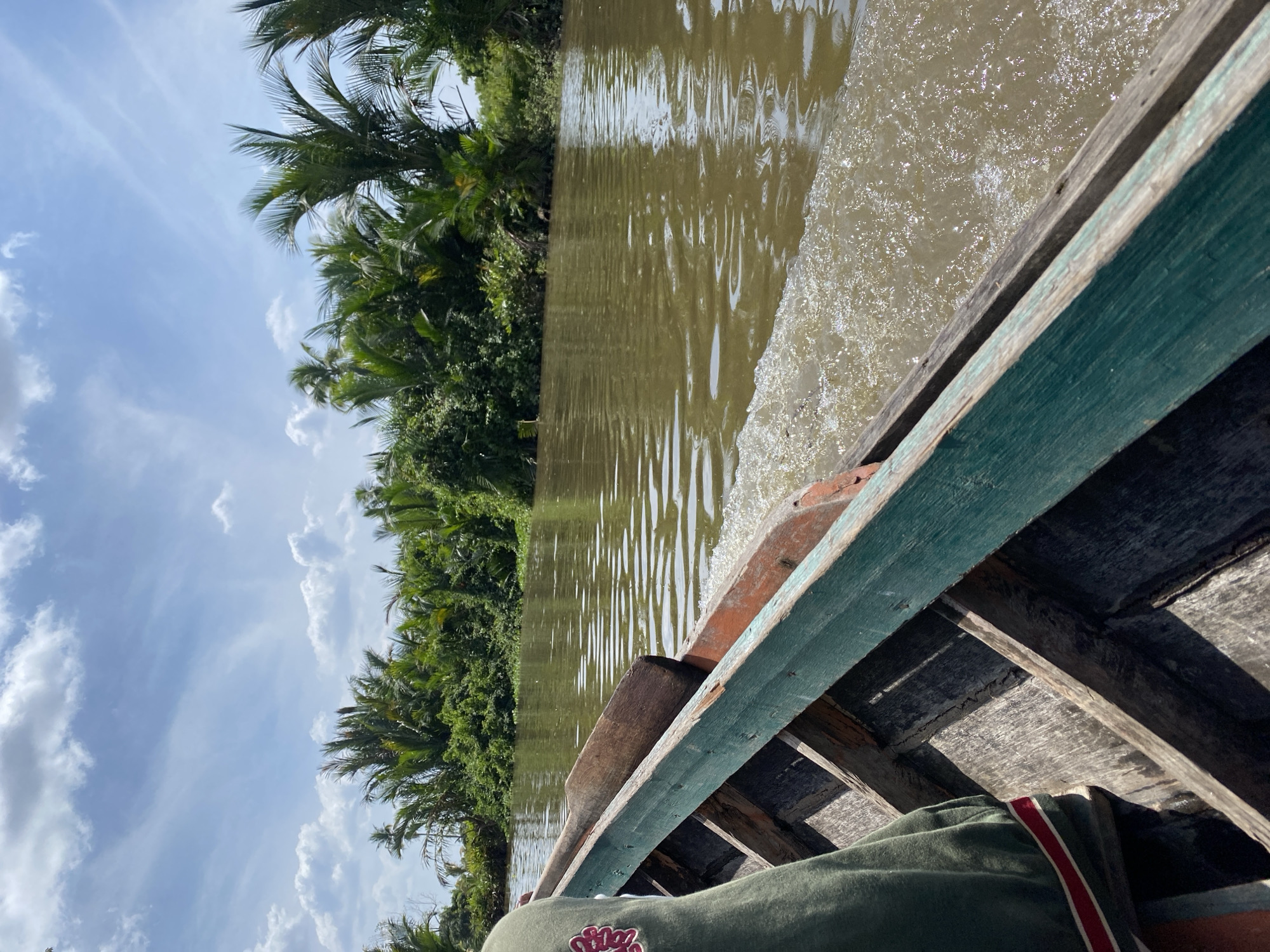 A first-person perspective shot from inside a wooden boat traveling down a muddy brown river. Lush green foliage and palm trees line the riverbanks under a partly cloudy blue sky. The boat has a weathered wooden interior and a green painted exterior edge. The water churns white from the boat's movement, creating small waves. A person wearing a green long-sleeved shirt with red and white stripes on the sleeve is visible on the left side of the frame. The scene is consistent with a boat trip on one of the many rivers in Banjarmasin, Indonesia, known for its riverine landscape and floating markets.