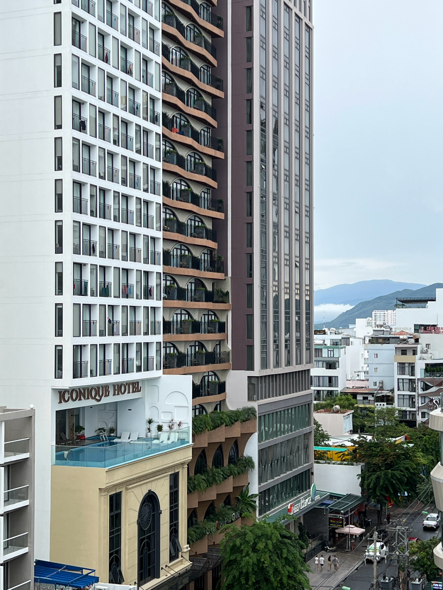 A view of the Iconique Hotel in Nha Trang, Vietnam, taken from a high vantage point. The hotel's distinctive white facade with numerous windows and balconies is visible on the left. Adjacent to it is a taller, darker building with a grid-like pattern of windows. In the foreground, a pool with lounge chairs sits atop a beige structure with arched windows, part of the hotel complex. The background shows a cityscape with other buildings and distant mountains, suggesting a dense urban environment. The sky is overcast. The street level below the hotel features trees, sidewalks, and a few vehicles and pedestrians.