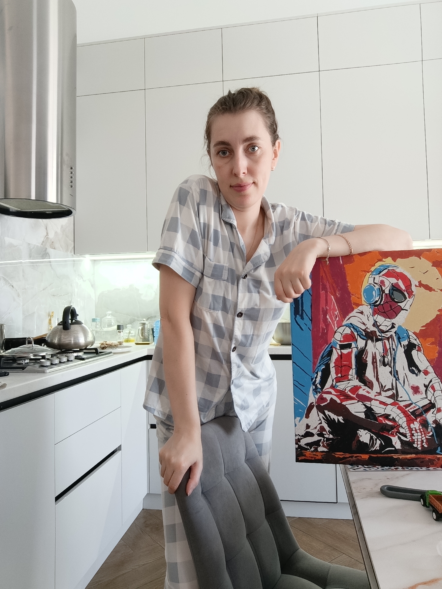A woman stands in a kitchen holding a painting. The kitchen has white cabinets and a marble backsplash. A kettle sits on a stovetop, and various items are arranged on the counter. The woman wears pajamas. The painting depicts a character resembling Spider-Man in a stylized, colorful manner. The location is implied to be in the city of Chaikovsky, Russia.