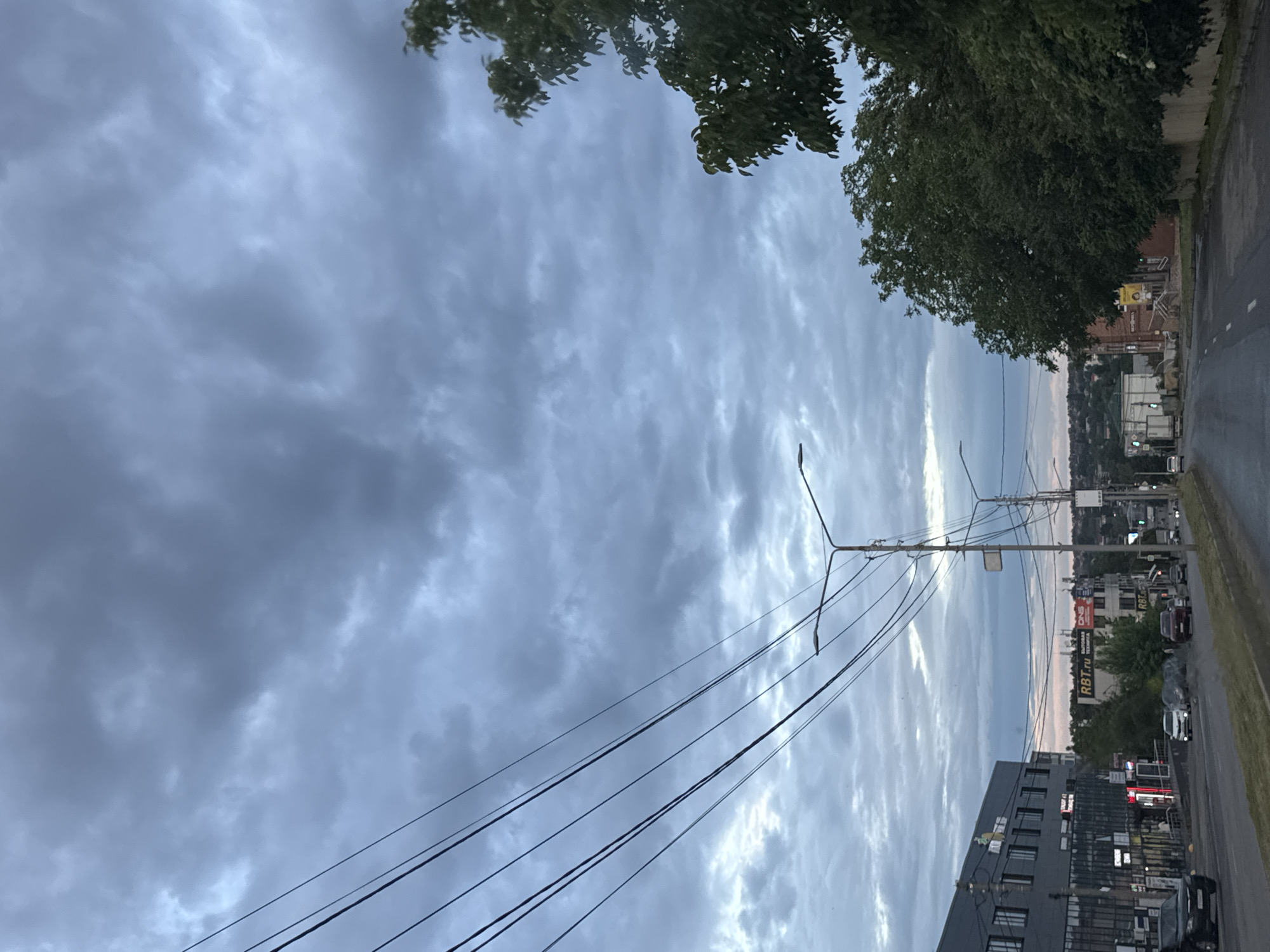 The image depicts a street scene in Mikhaylovsk, Russia, under a cloudy sky. The perspective is from a vehicle traveling on a paved road. On the left side of the road, there is a modern, dark-colored building with multiple windows and a commercial facade. Signs such as "RBТ.RU" are visible on other buildings further down the street. Power lines stretch across the upper portion of the frame. On the right side of the road, several trees with dense green foliage are present. Traffic lights, streetlights, and various vehicles are visible on the road. The overall atmosphere suggests a somewhat overcast day in an urban or semi-urban environment. There are no visible people in the image.