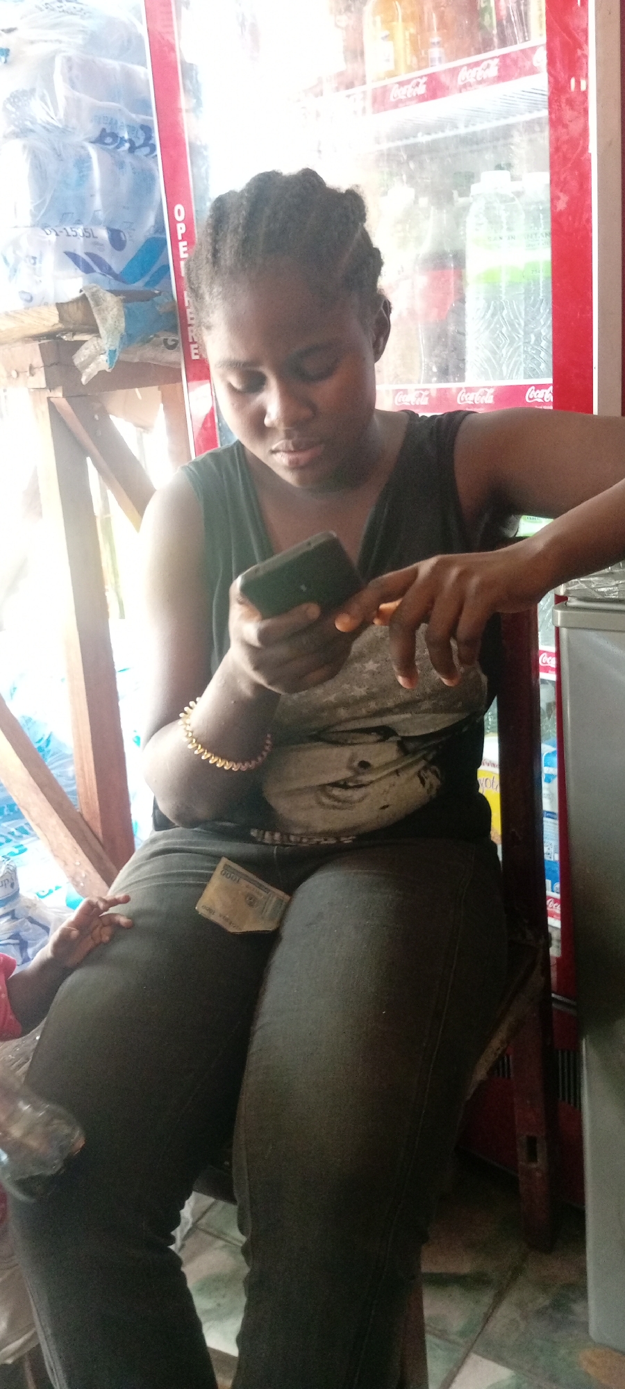 A woman is seated indoors, likely in a small store in Kwachiri, Nigeria. She's focused on a mobile phone she holds in her hands. She wears a dark tank top, jeans, and a gold bracelet. A banknote is visible on her lap. A refrigeration unit filled with Coca-Cola and bottled water is behind her. Plastic-wrapped water bottles are stacked to her left.